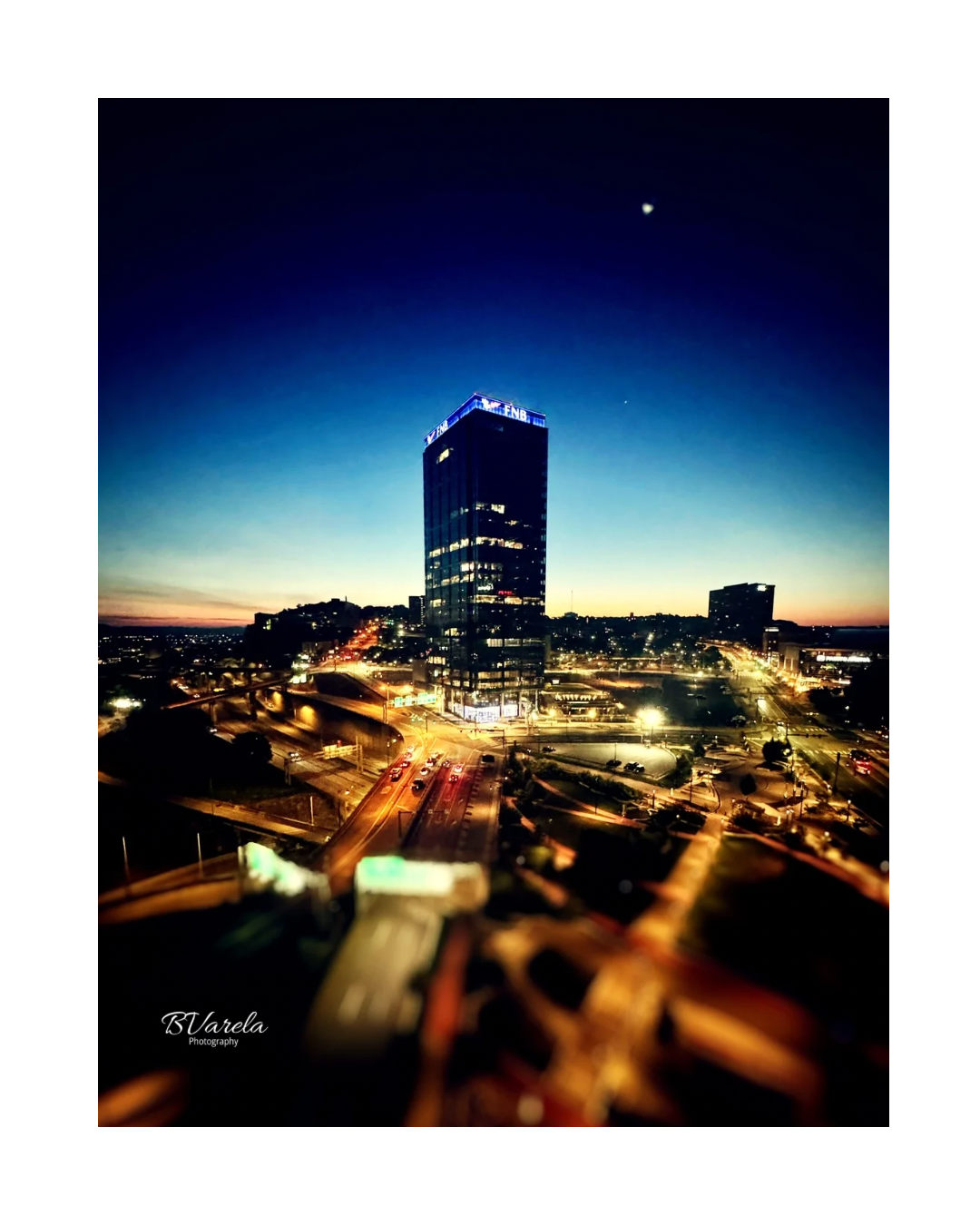 Aerial night cityscape with illuminated skyscraper, glowing streets, and crescent moon in deep blue twilight sky.