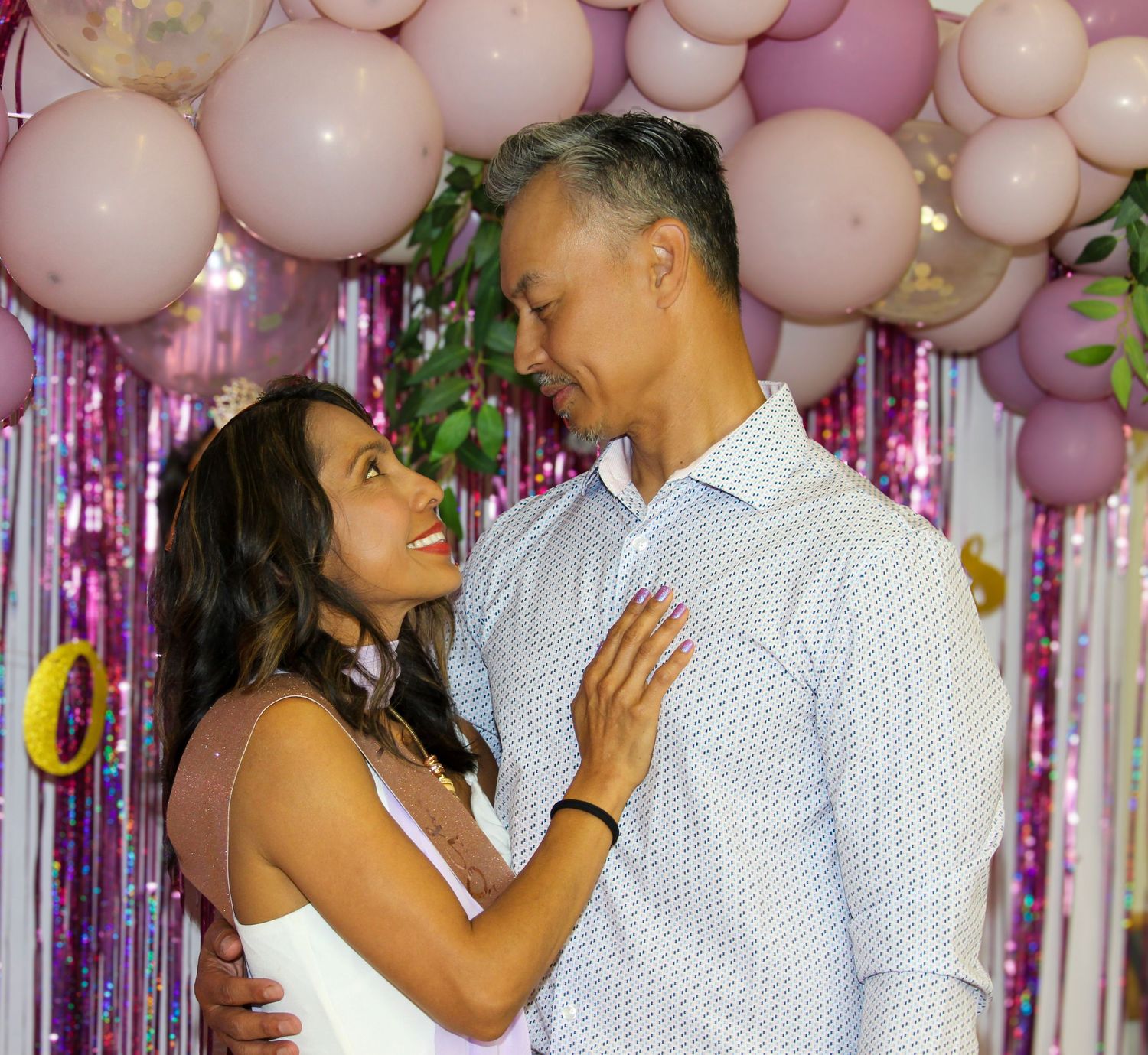 A couple smiles at each other at a pink balloon and tinsel backdrop celebration event.