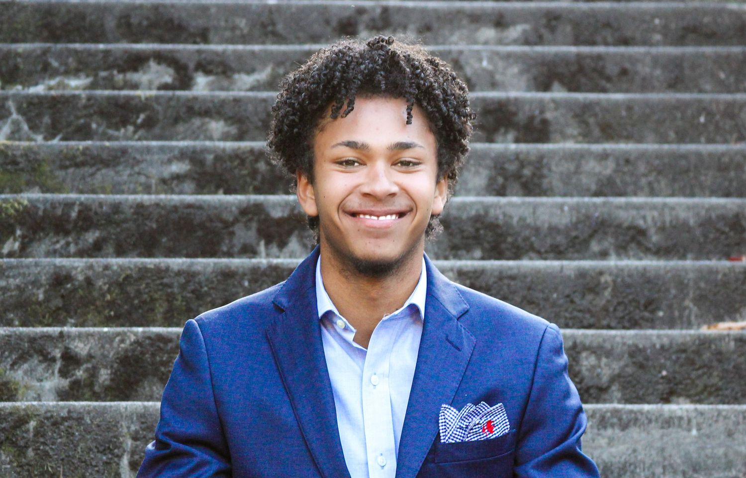 Young man in blue blazer smiling confidently on stone steps outdoors.