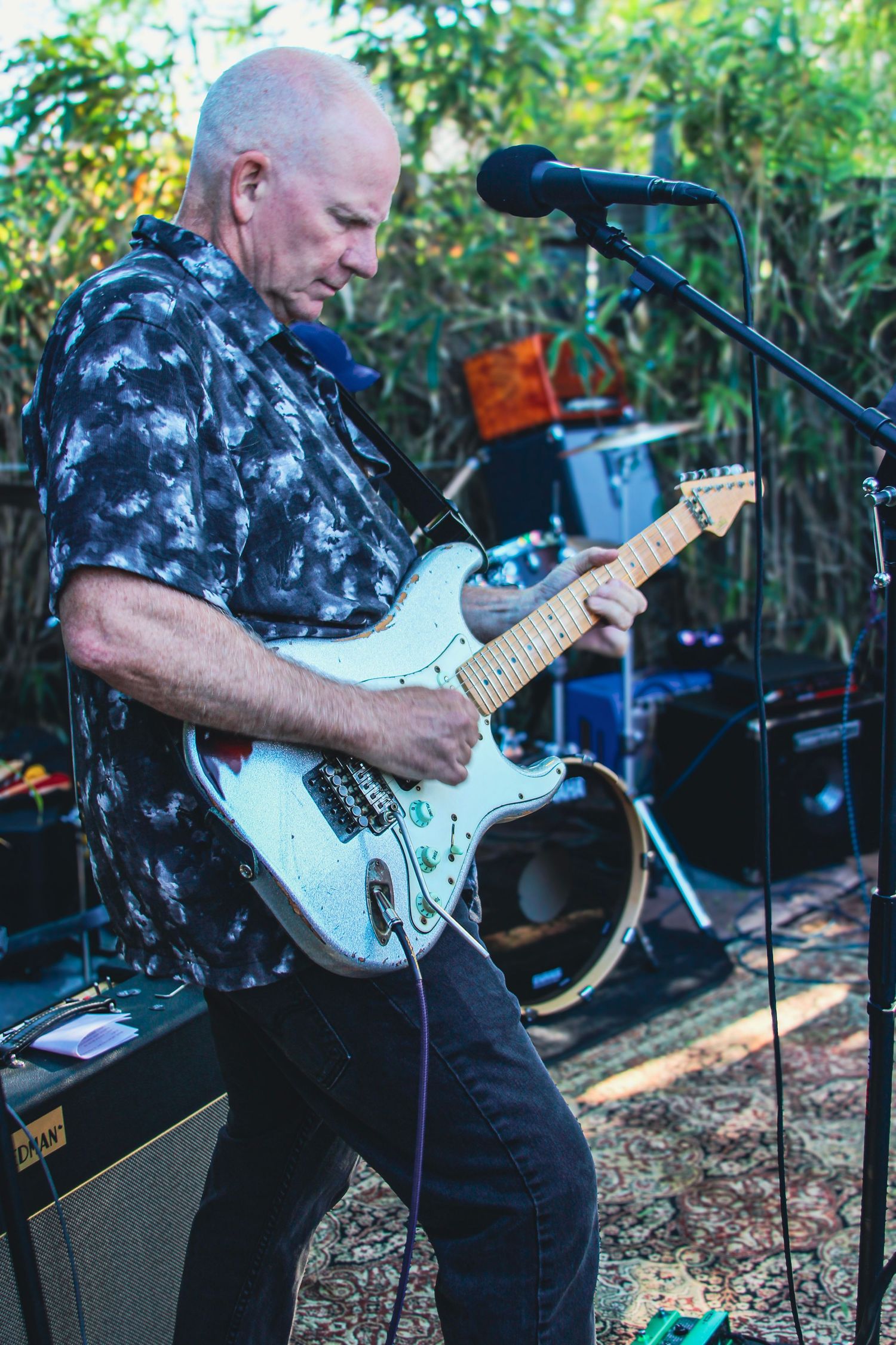 Bald guitarist playing white Stratocaster electric guitar on outdoor stage with microphone stand nearby.