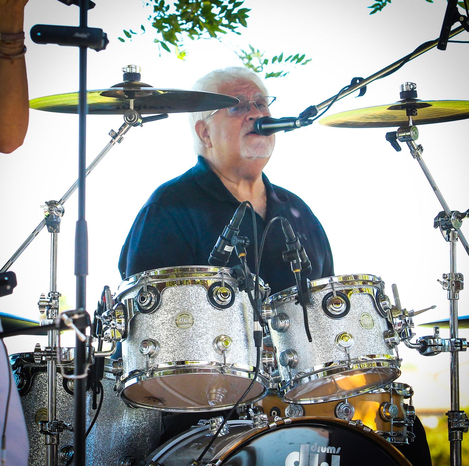 A drummer with a mustache performs on a silver drum kit at an outdoor music event.