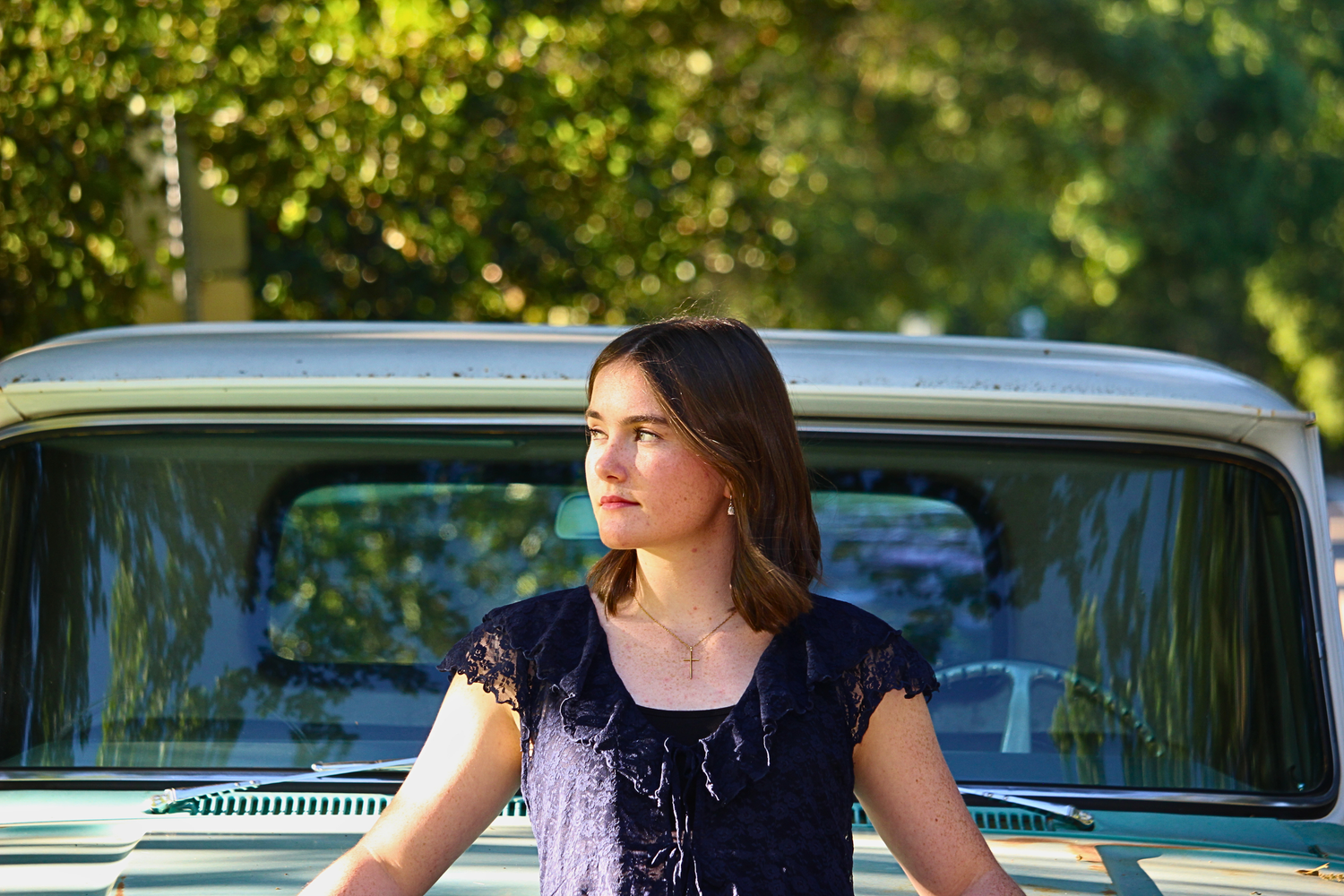 Young woman in dark lace top leaning against a vintage truck outdoors with green trees in background.