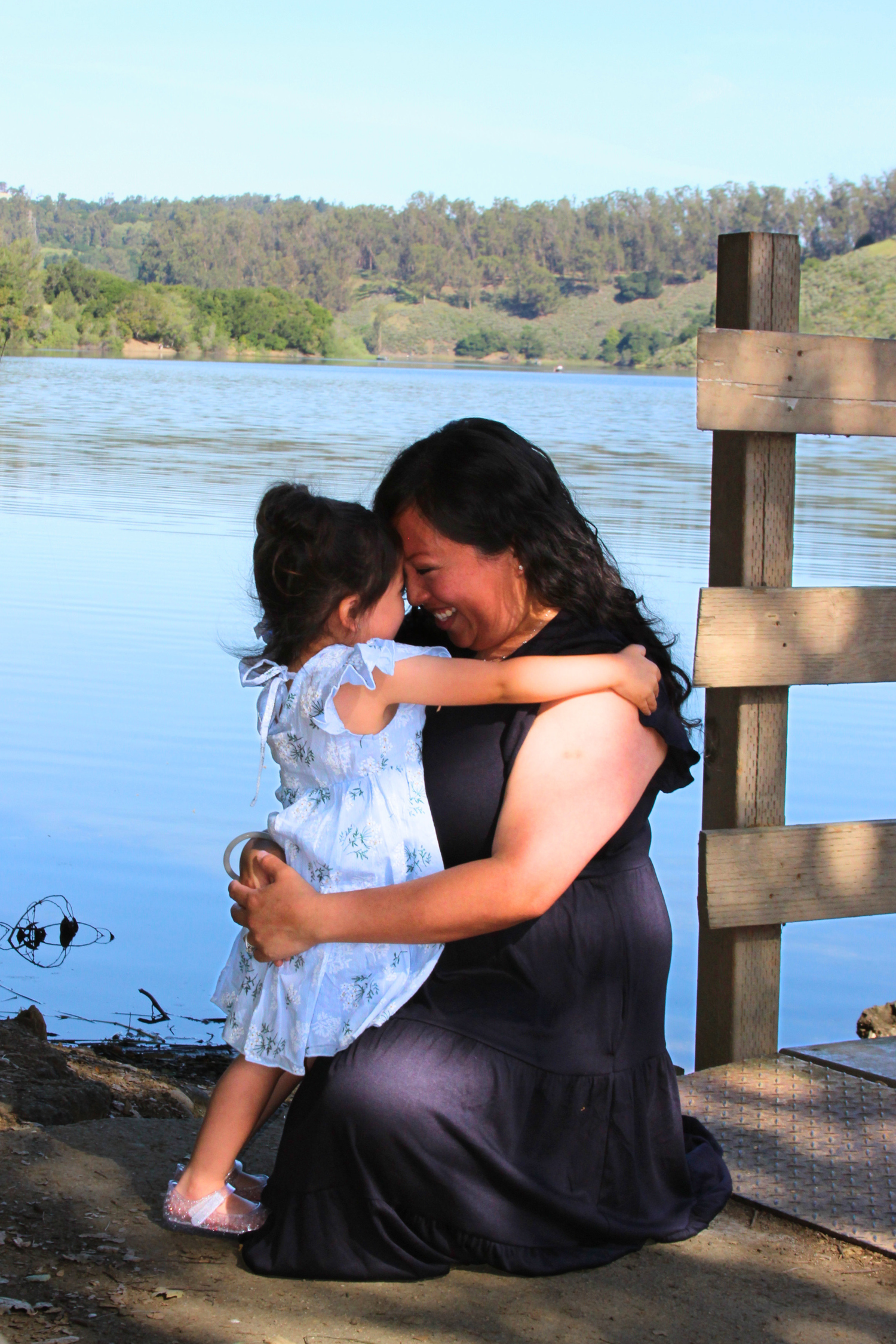 A woman kneels by a lakeside dock, lovingly embracing a young girl in a floral dress on a sunny day.