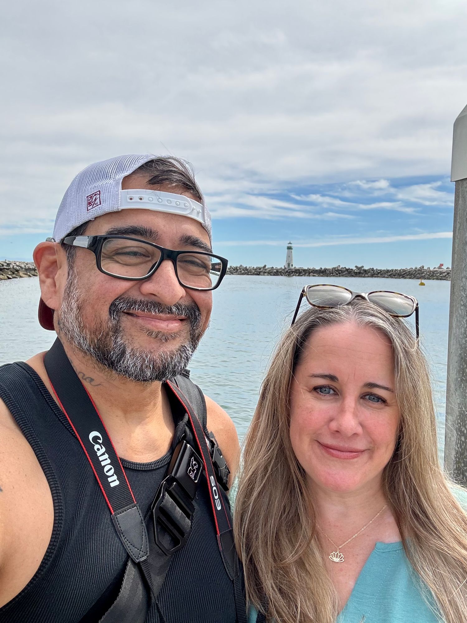 A man and woman take a selfie near a waterfront with a lighthouse and cloudy sky in the background.