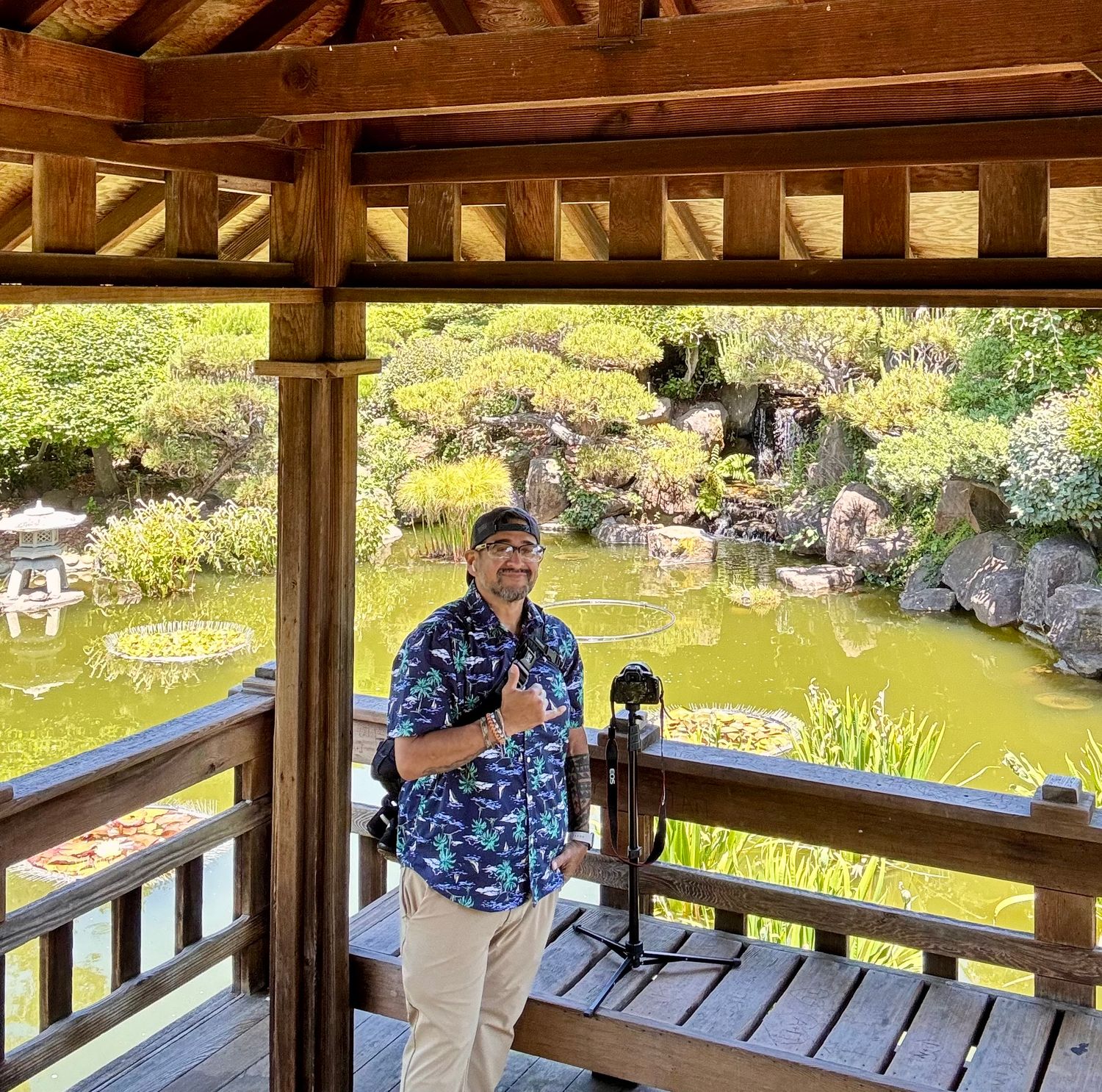Man in floral shirt with camera gear stands in Japanese garden pavilion overlooking serene koi pond.