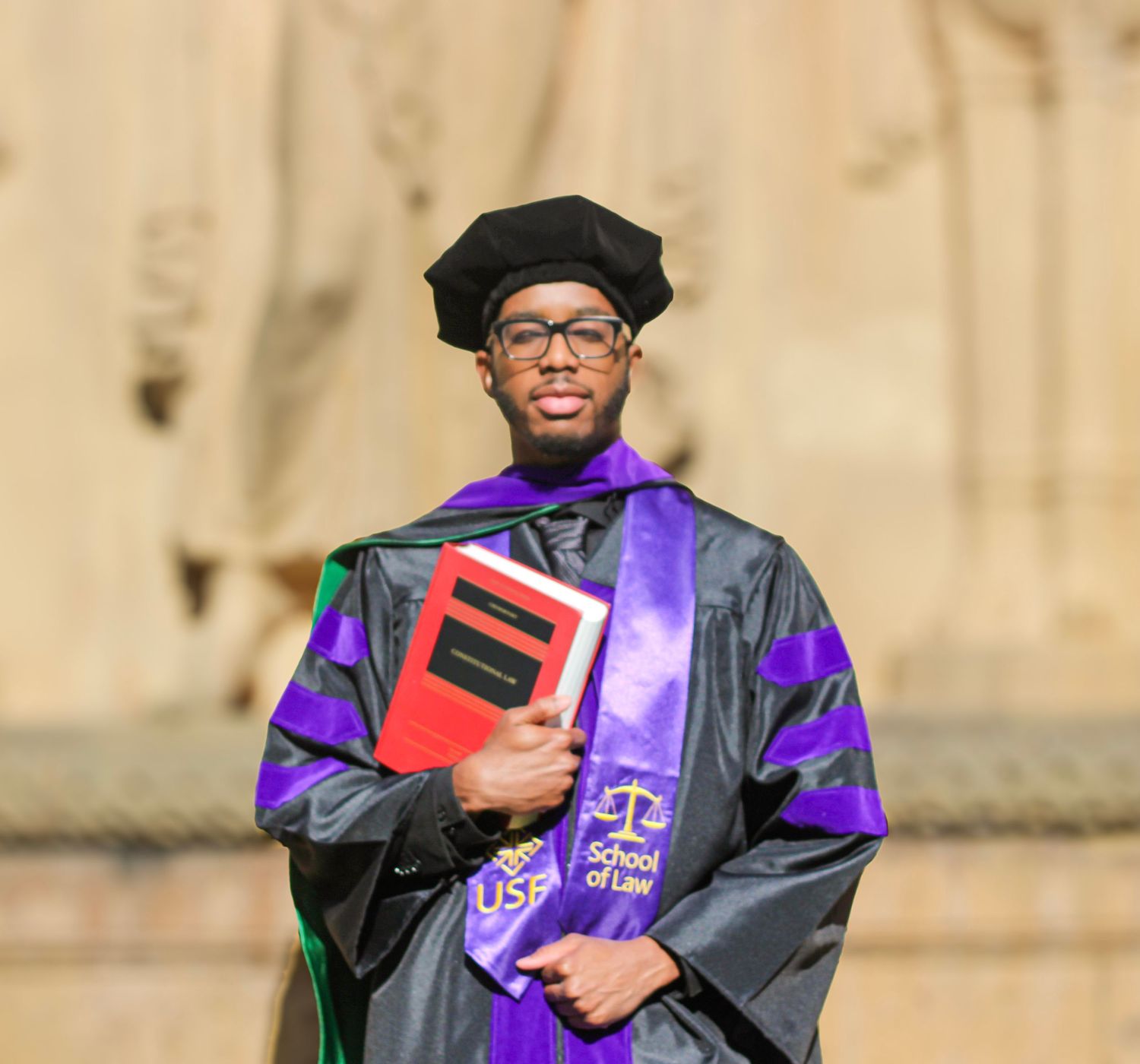 Graduate in black and purple law school regalia holds books at outdoor commencement ceremony.