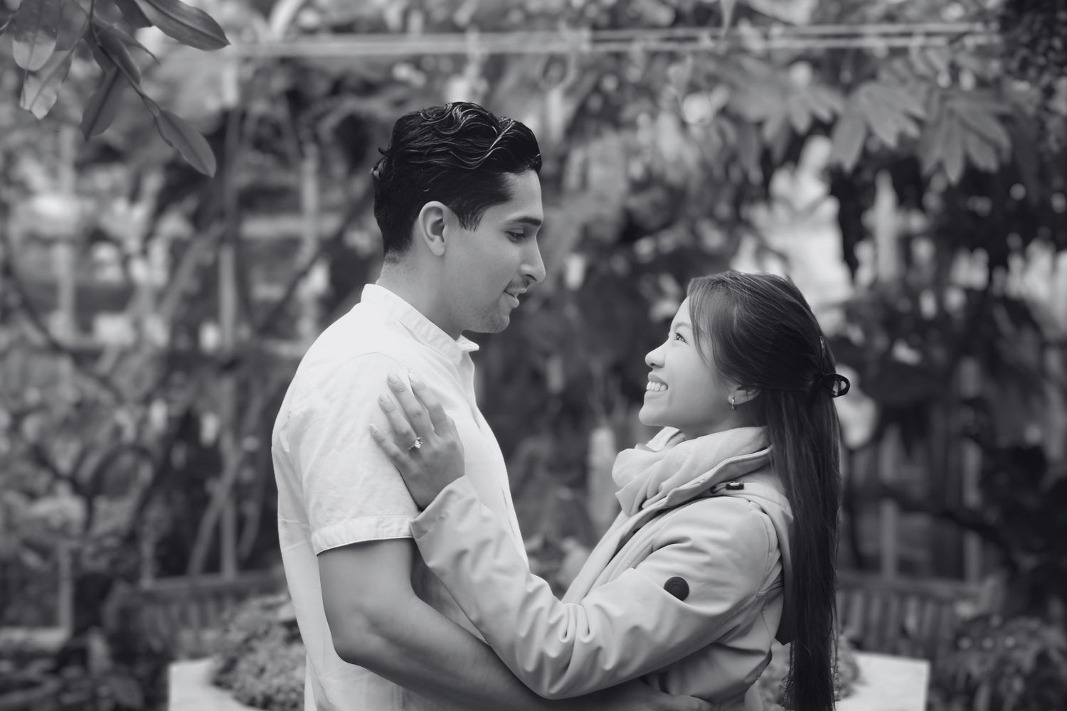 Black and white photo of a romantic couple embracing and gazing at each other in a lush garden setting.