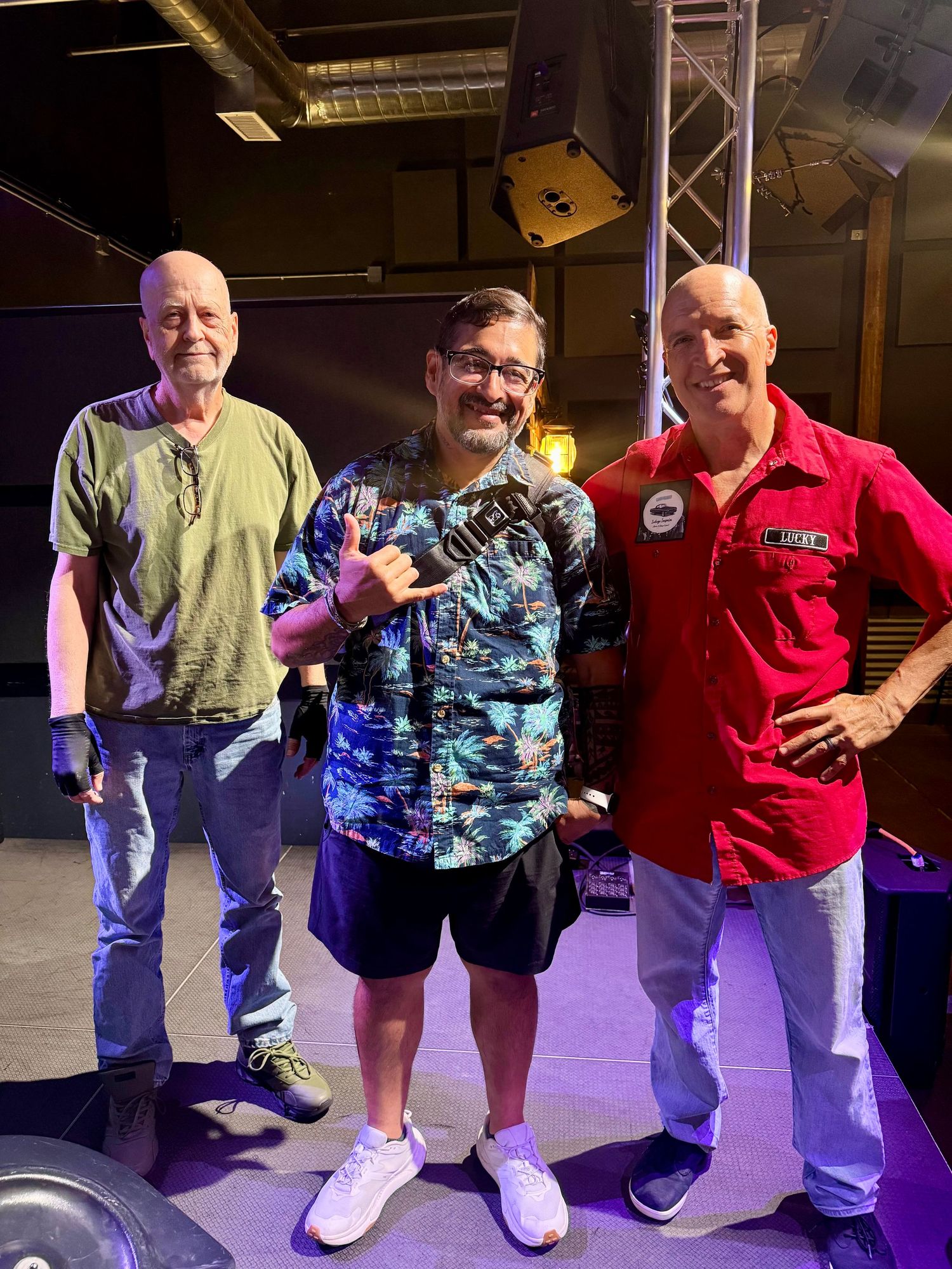 Three men posing together indoors on a stage, one wearing a colorful Hawaiian shirt making a hand gesture.