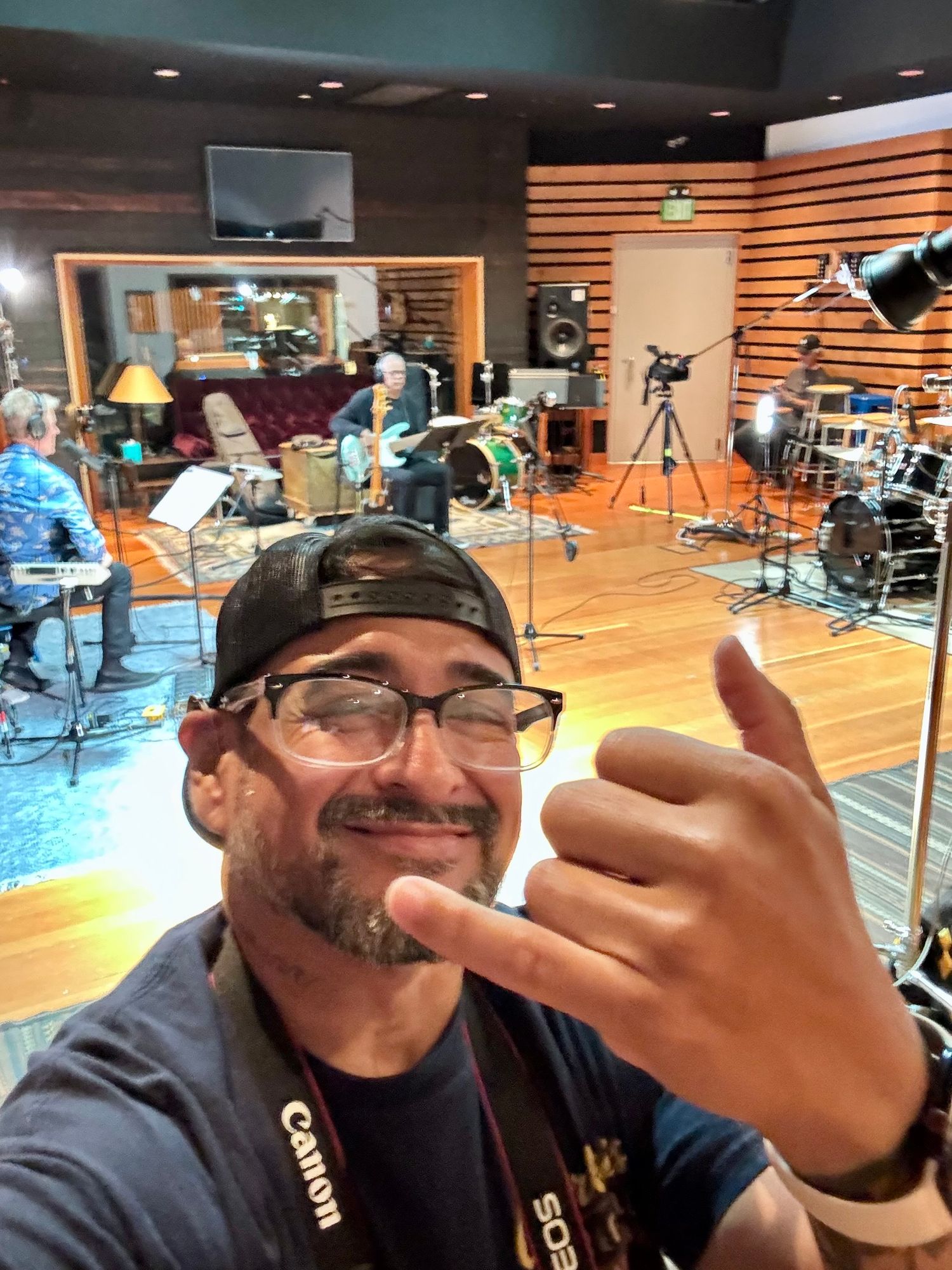 Man taking selfie in recording studio with Canon camera strap, giving shaka sign, musicians playing instruments in background.