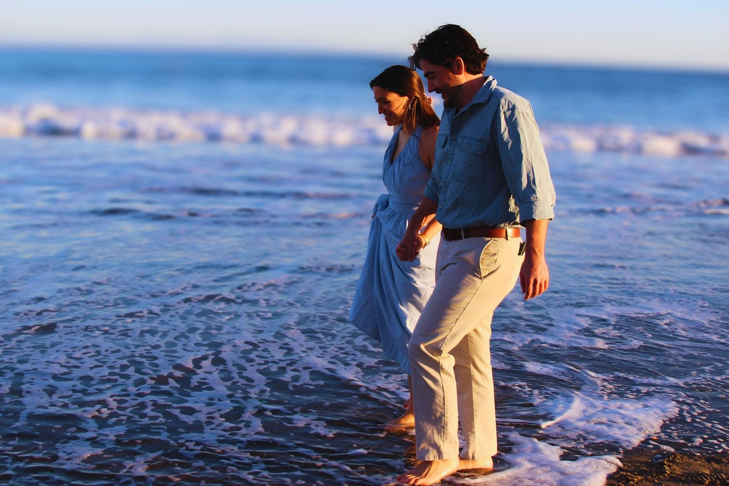 Couple walking barefoot on a sandy beach at sunset, waves gently washing over their feet.
