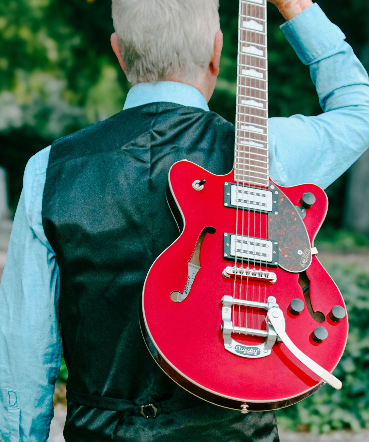 Man in vest holding red Gretsch semi-hollow electric guitar outdoors with green foliage background.