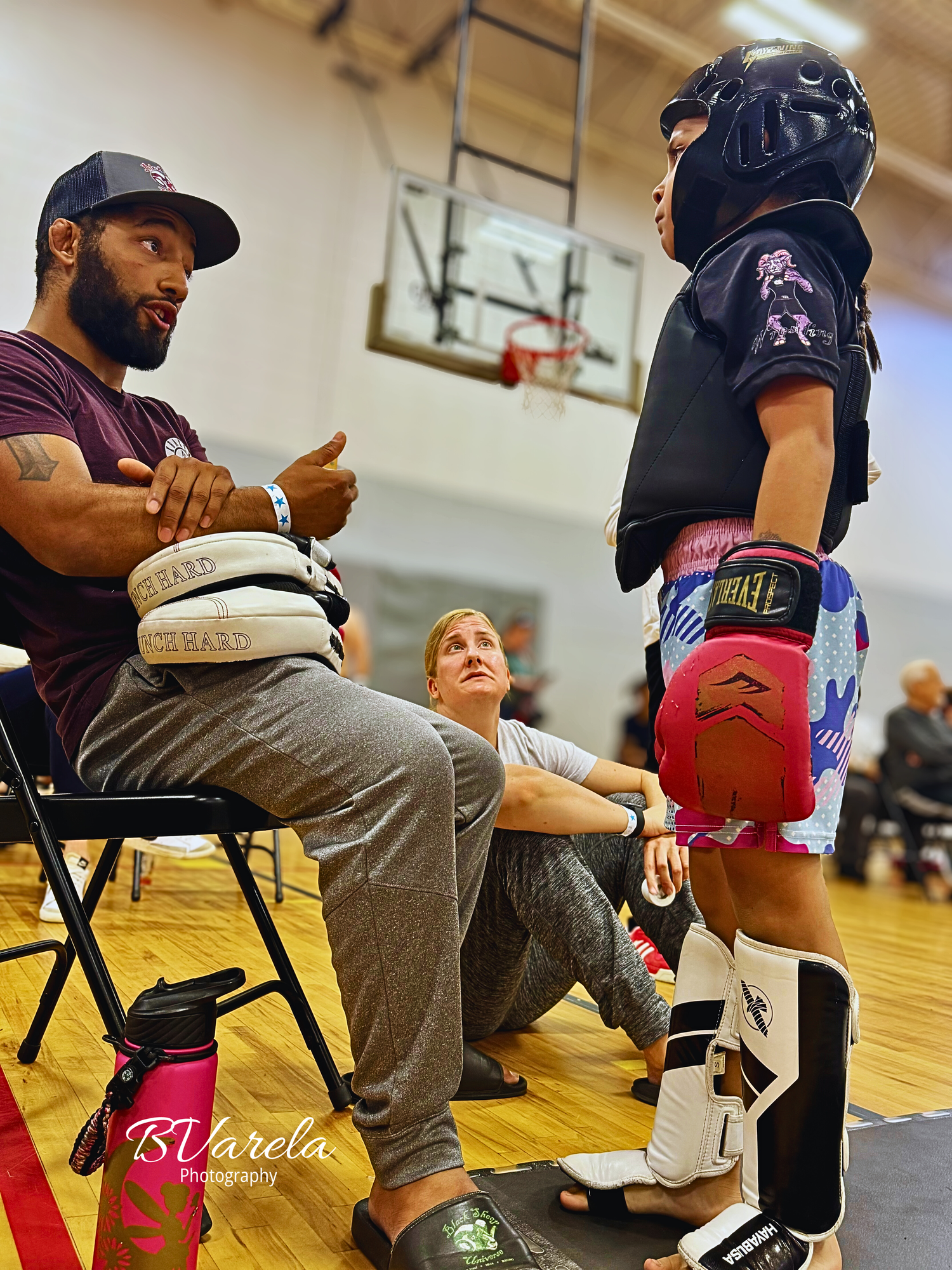A martial arts coach speaks with a young fighter in protective gear at an indoor gym during a kickboxing tournament.