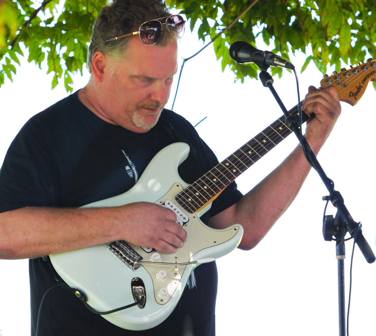 Man in black t-shirt playing white Stratocaster electric guitar outdoors at a live music event near a microphone stand.