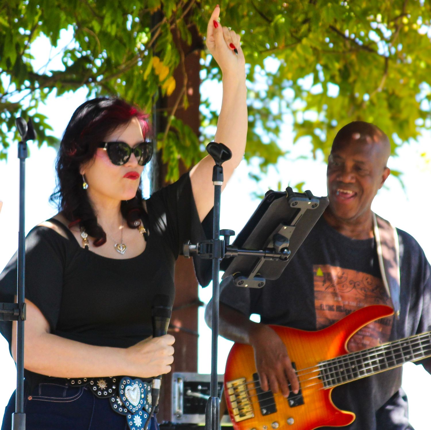 Woman singer with raised fist and guitarist performing at outdoor concert under leafy trees.
