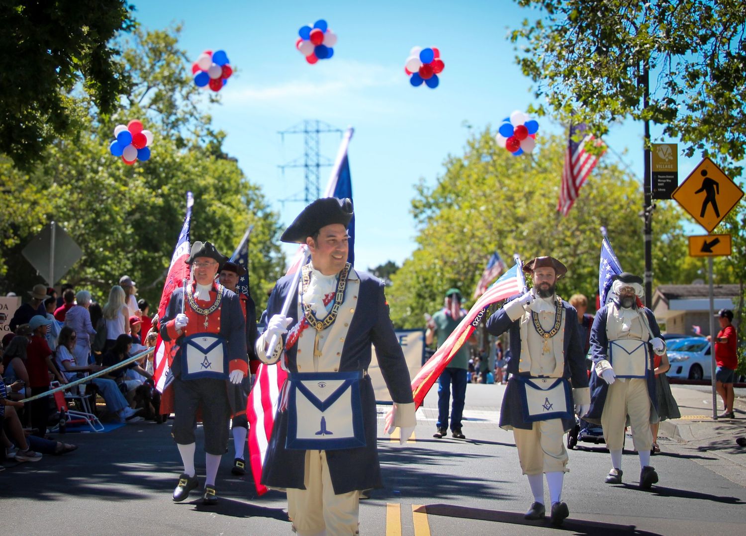 Freemasons in colonial costumes march in a Fourth of July parade with American flags and colorful balloons.