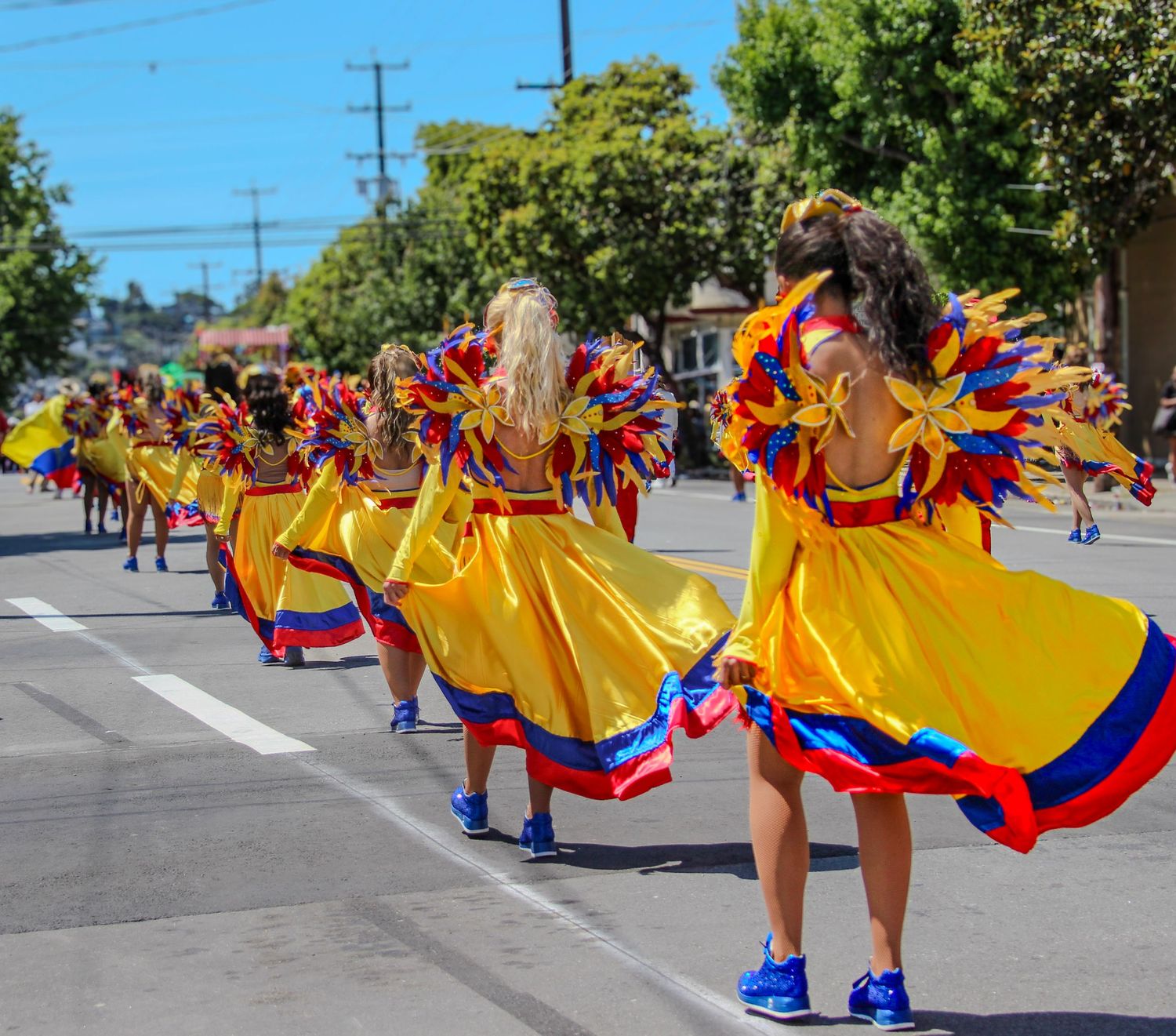 Dancers in yellow Colombian dresses with colorful feathered costumes perform in a vibrant street parade on a sunny day.