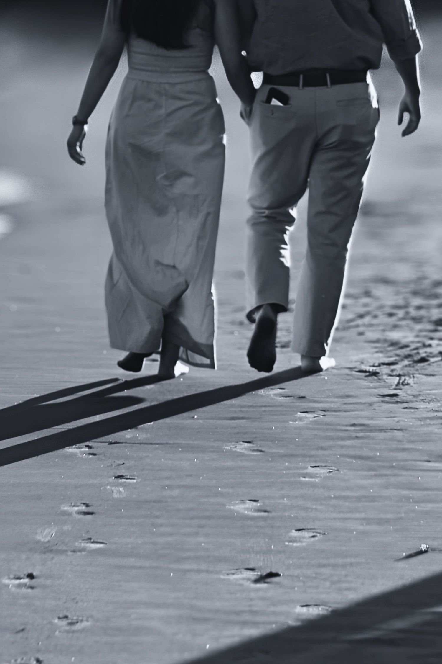 Black and white photo of a couple walking hand in hand on a sandy beach, leaving footprints behind them.