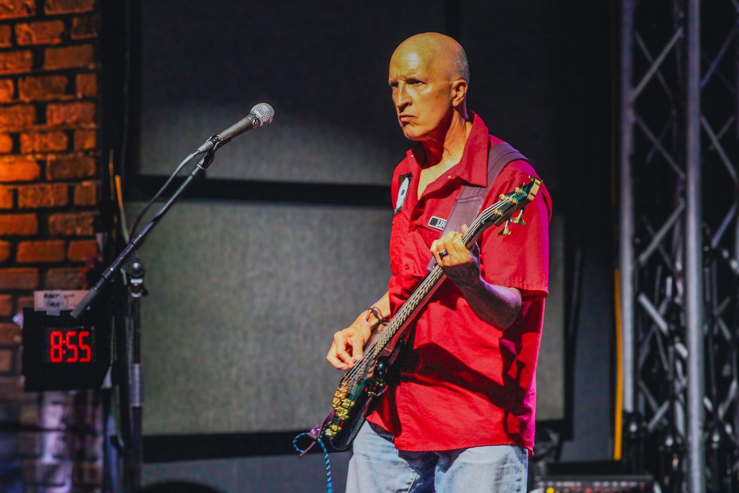 Bald guitarist in red shirt performs on stage with electric guitar near microphone stand.
