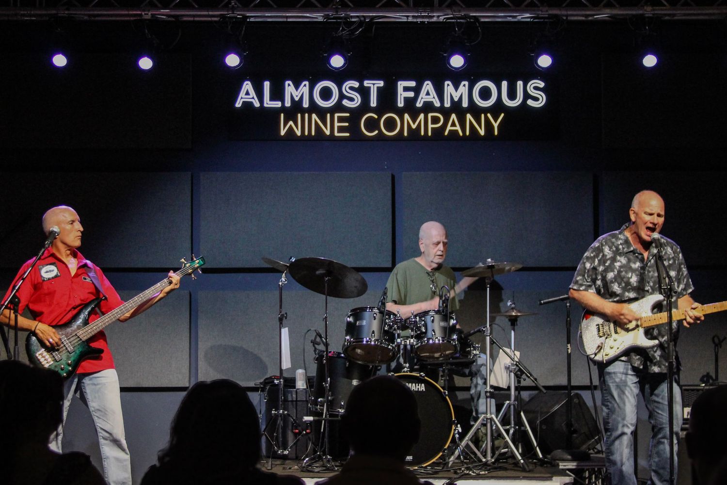 Three musicians perform live at Almost Famous Wine Company, featuring a bassist, drummer, and guitarist on a dimly lit stage.