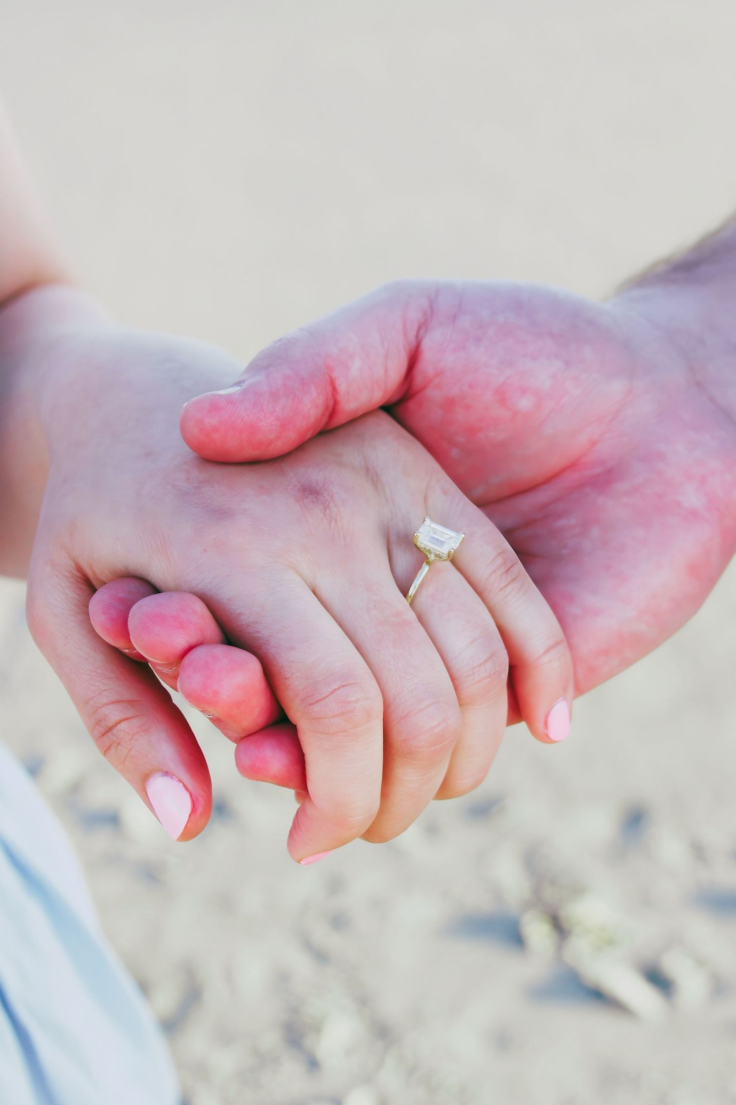 Close-up of couple holding hands on sandy beach, woman wearing elegant emerald-cut diamond engagement ring with pink nails.