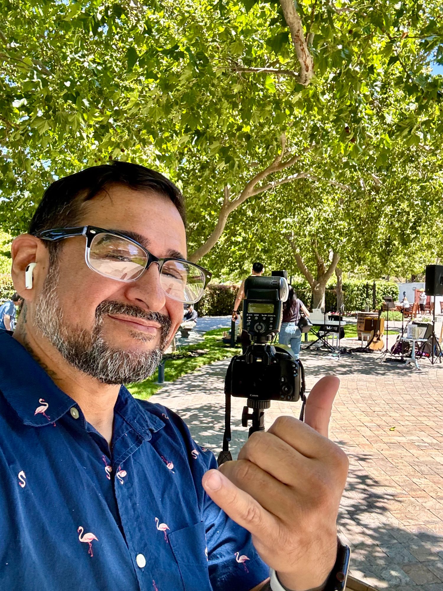 Man with glasses giving thumbs up at outdoor market with camera on tripod under green trees.