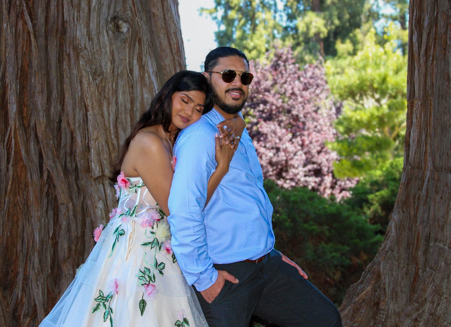 Couple posing between redwood trees, woman in floral dress leaning on man in blue shirt, cherry blossoms in background.