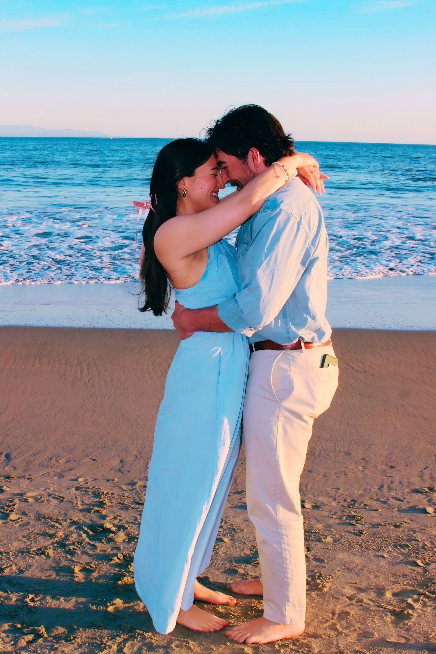 Romantic couple embracing and almost kissing on a sandy beach at sunset with ocean waves in the background.