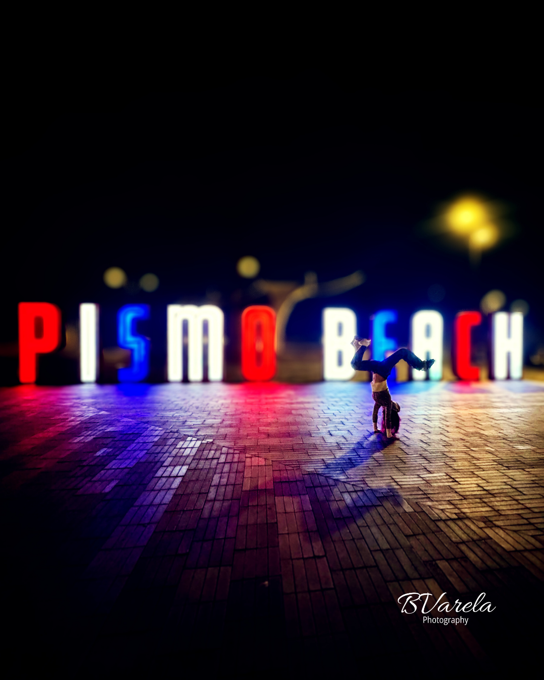 Person doing a handstand in front of illuminated Pismo Beach sign at night. pismo beach