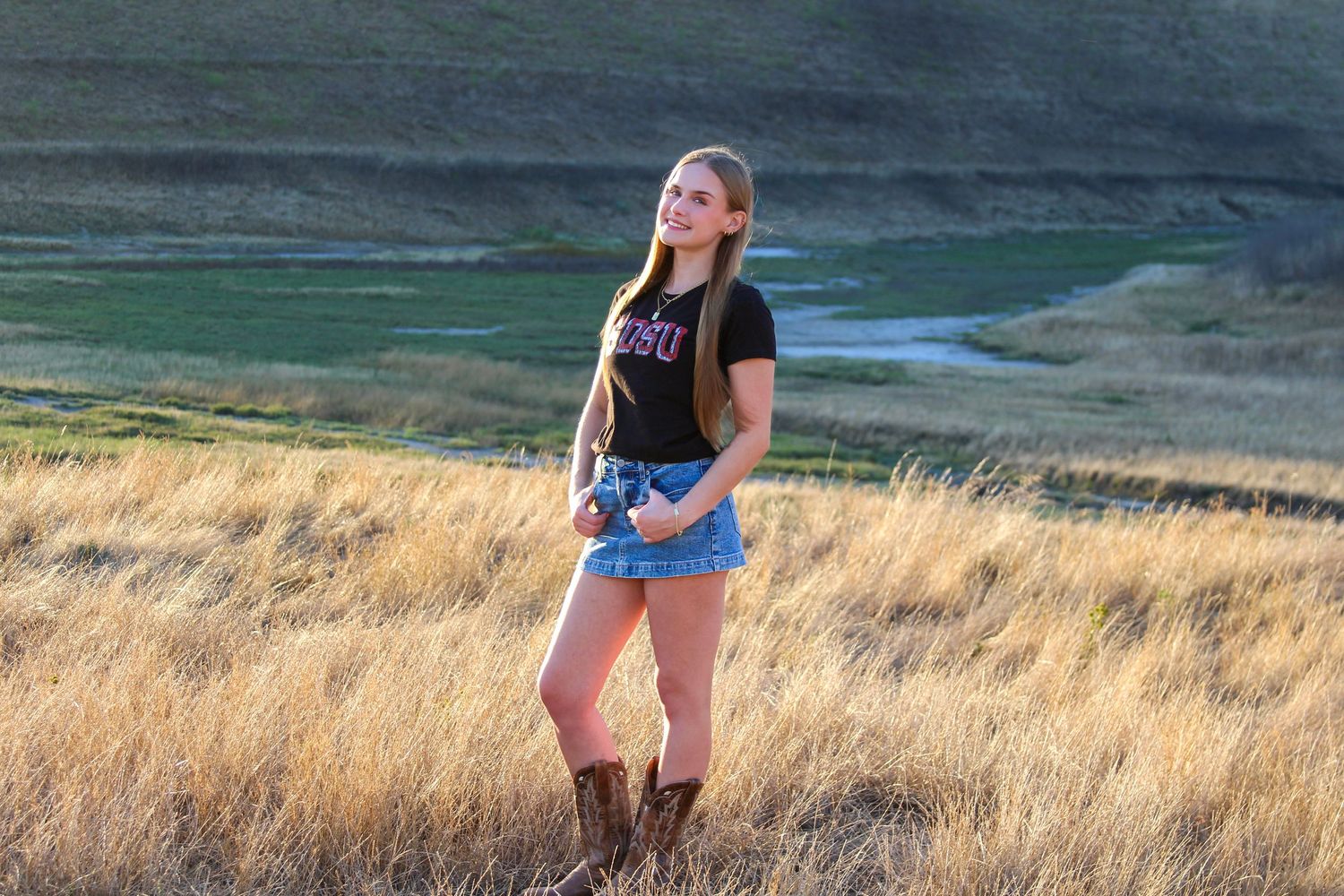 Young woman in black tee and denim shorts standing in golden grassland with scenic valley backdrop.