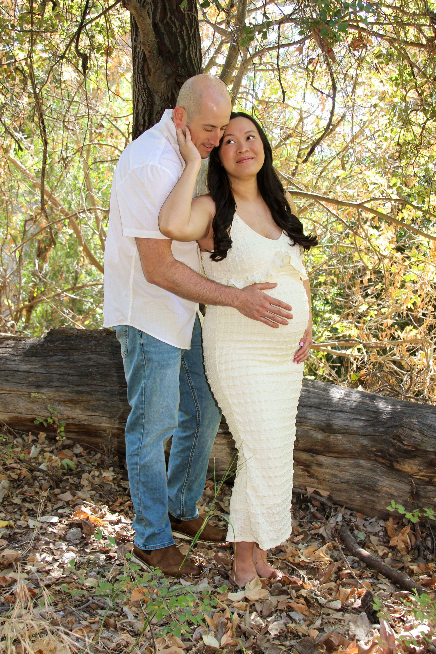 Couple sharing a tender moment in autumn woods, pregnant woman in cream dress cradled by bald man in white shirt.
