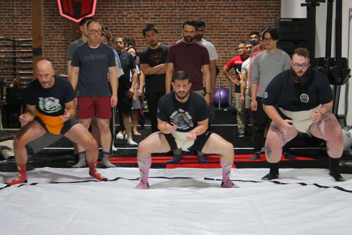 Three men compete in a sumo wrestling event at a gym, watched by a crowd of spectators standing behind them.