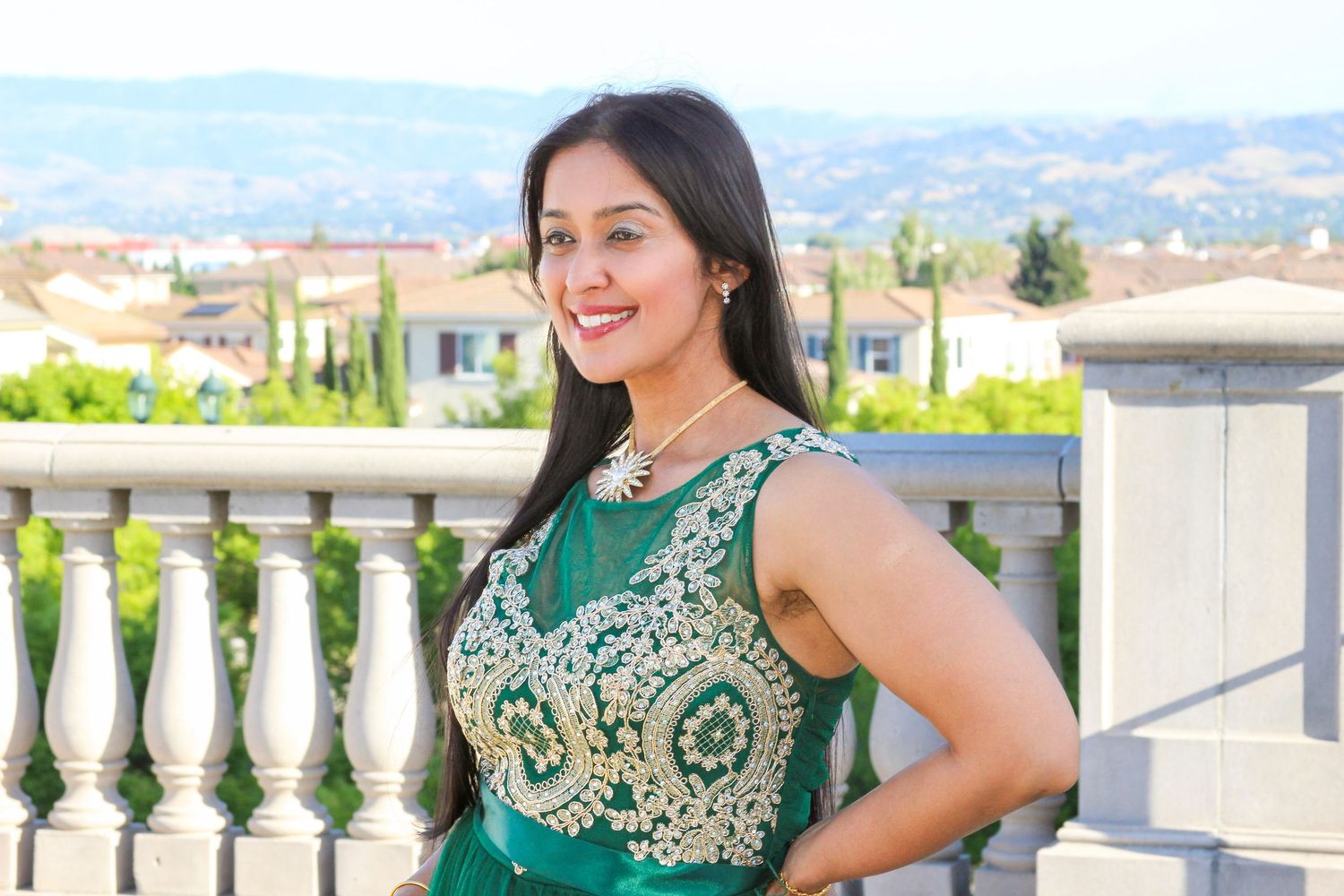A woman in an elegant green and gold embroidered dress smiles outdoors near a decorative balustrade with hills behind her.
