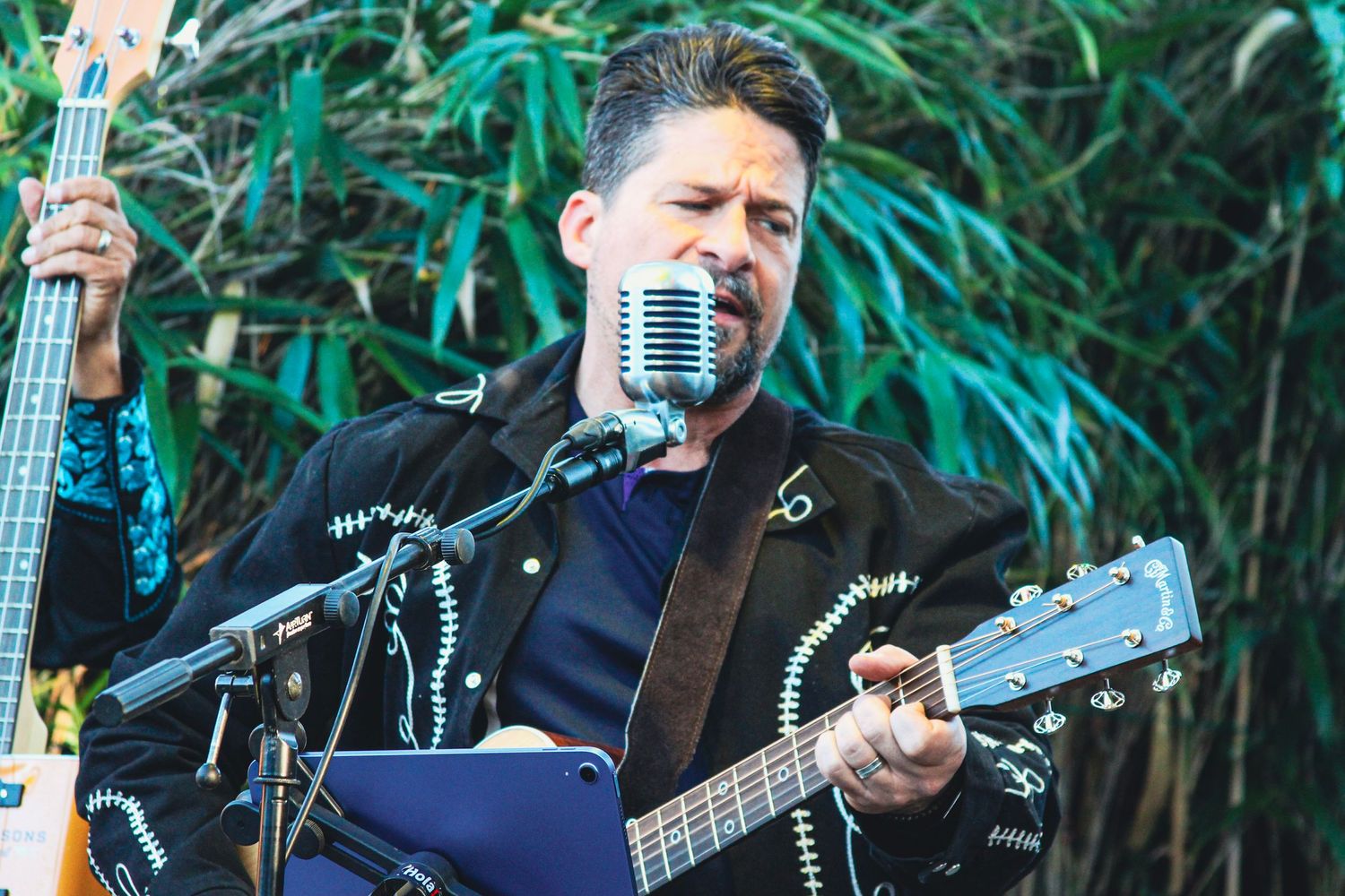 Male guitarist in black mariachi jacket performs at outdoor venue with vintage microphone and lush green foliage backdrop.