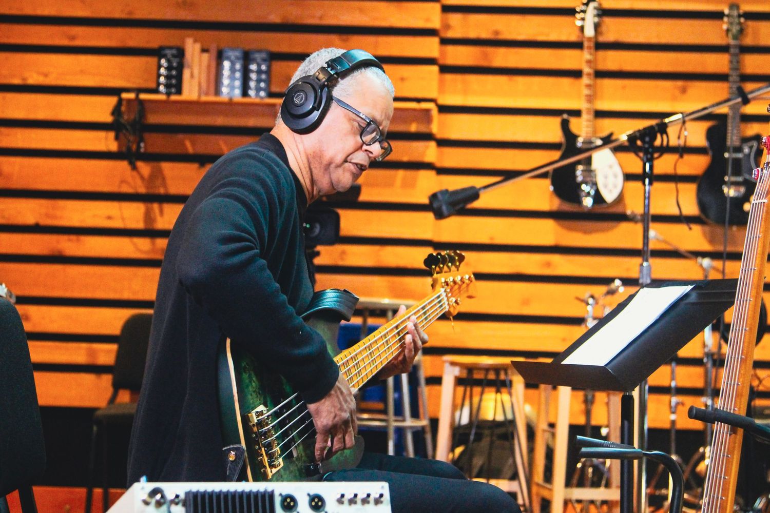 Musician wearing headphones plays bass guitar in a recording studio with instruments on the wall.