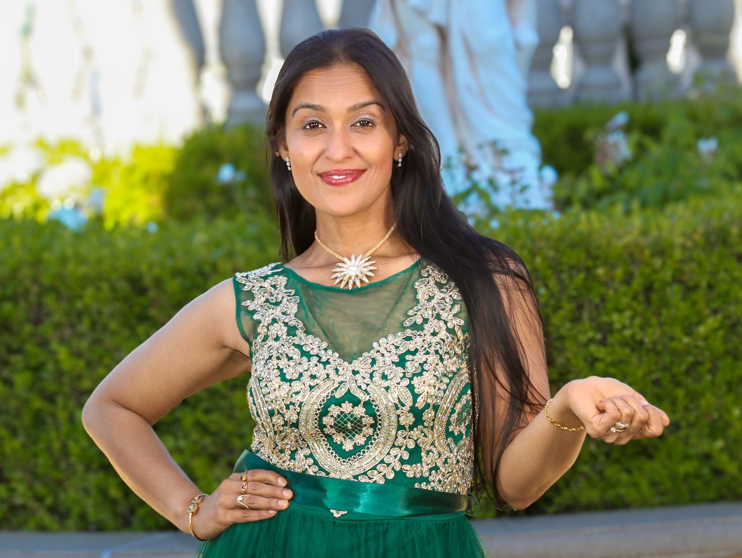 Woman in elegant green embroidered dress with floral necklace posing outdoors near lush hedges.