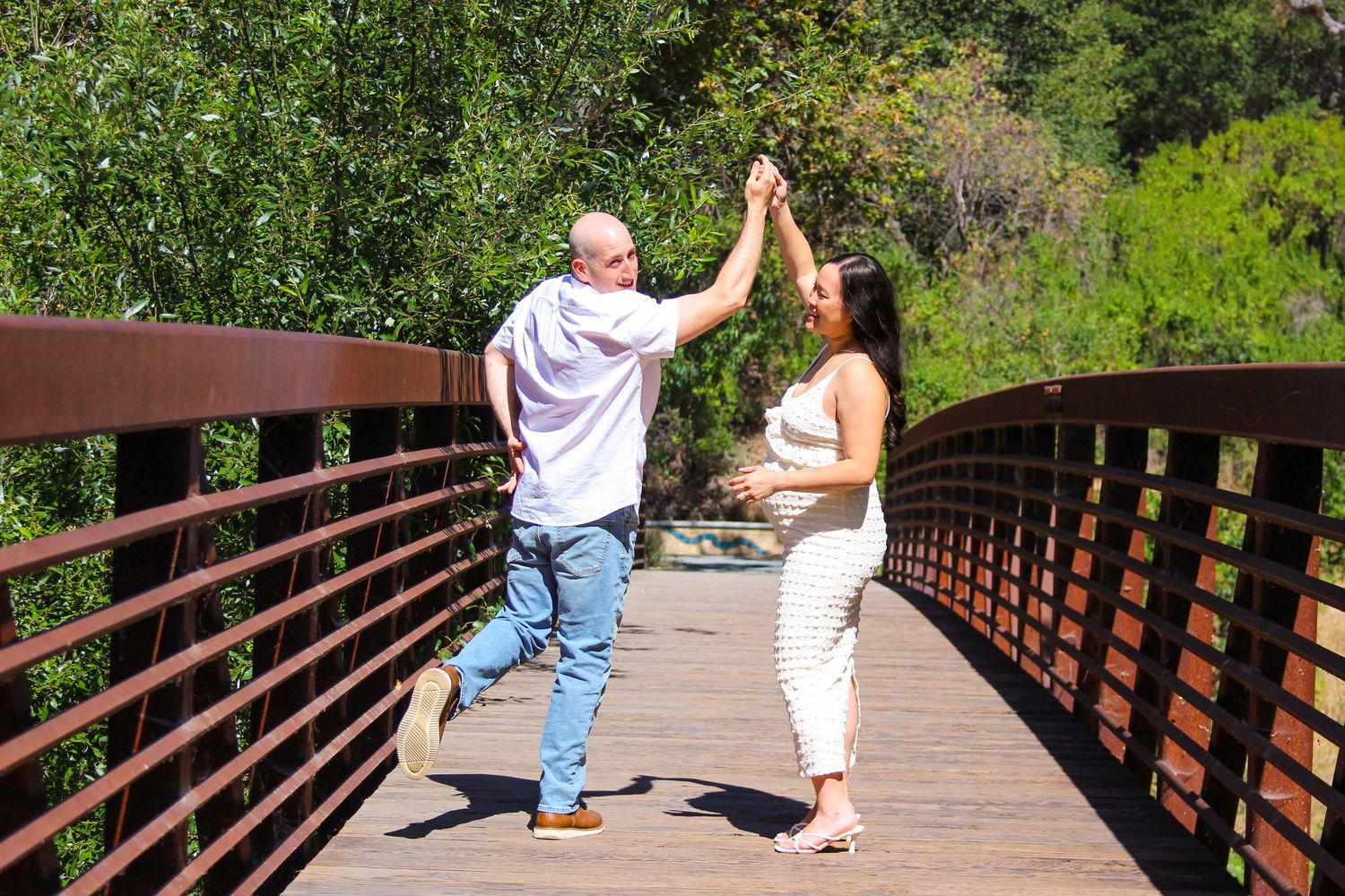 Pregnant couple joyfully high-fiving on a wooden bridge surrounded by lush green trees on a sunny day.