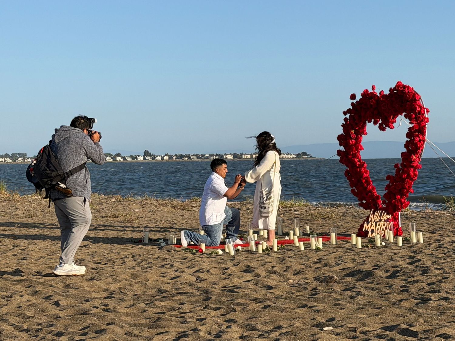 Man proposes on beach with red heart balloon arch, candles, and photographer capturing the romantic moment.