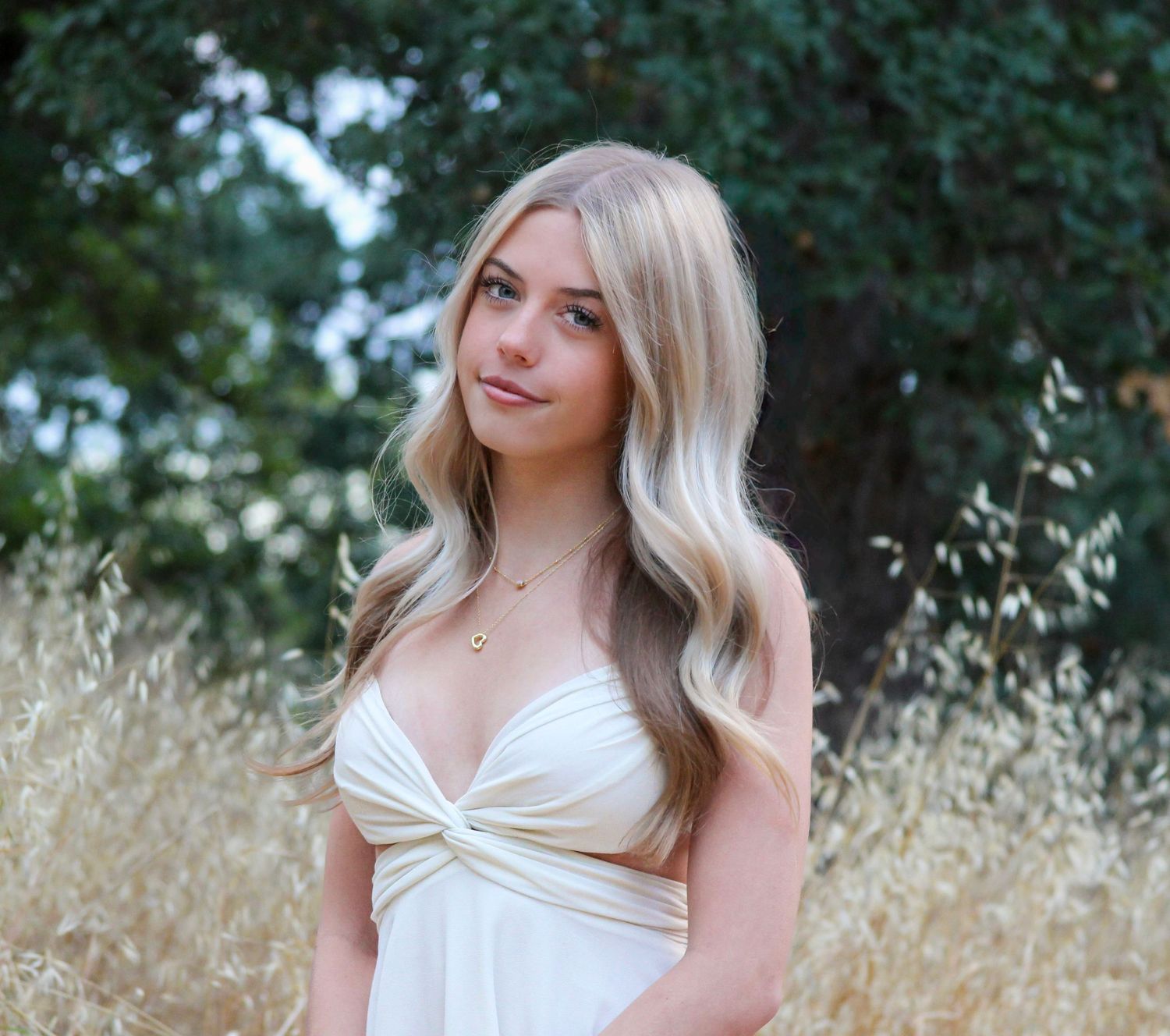 Young blonde woman in white dress posing in golden field with trees, long wavy hair, delicate necklace, natural light.