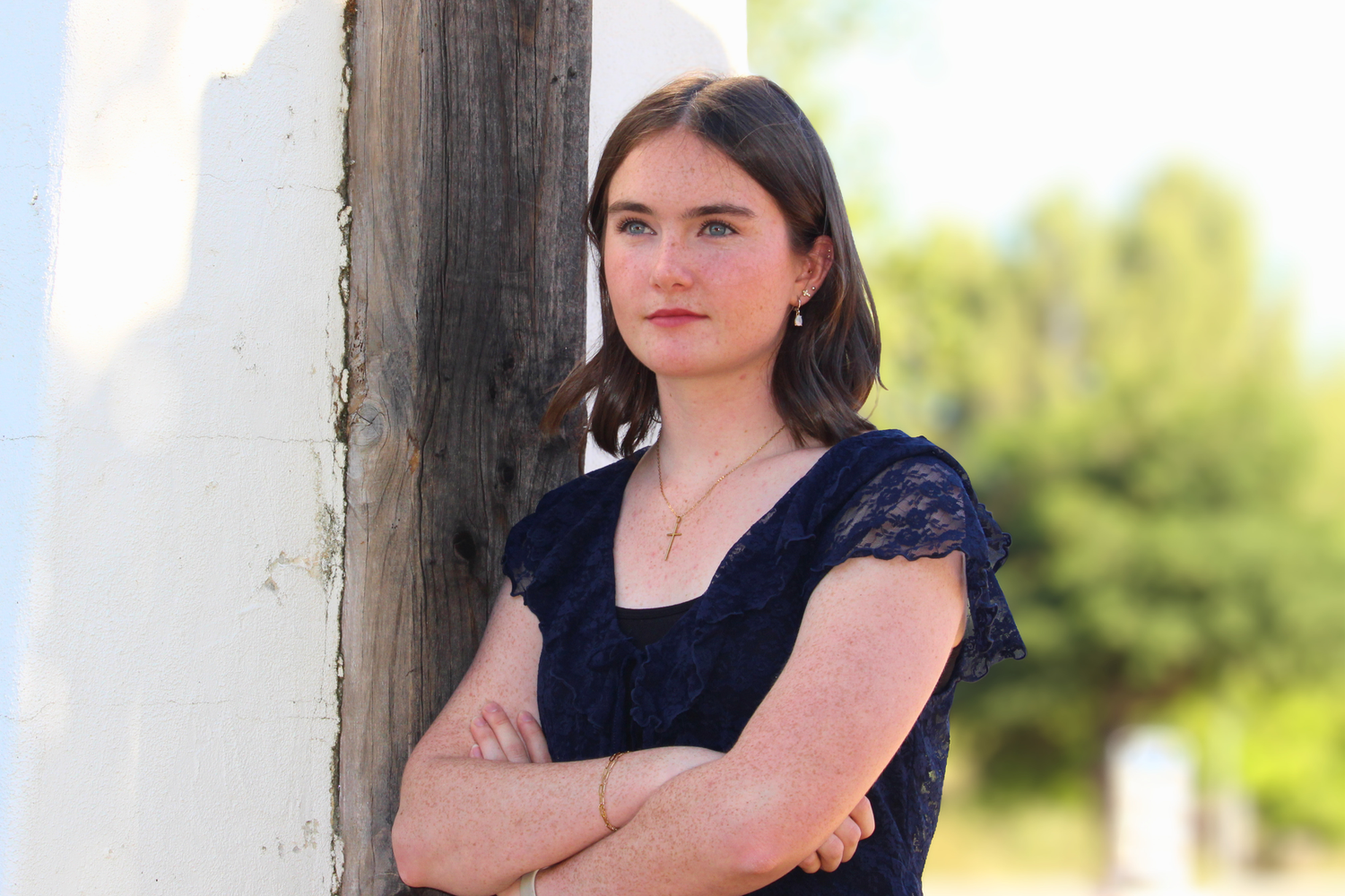Young woman in navy lace top leaning against wooden post with arms crossed, outdoor portrait photography.