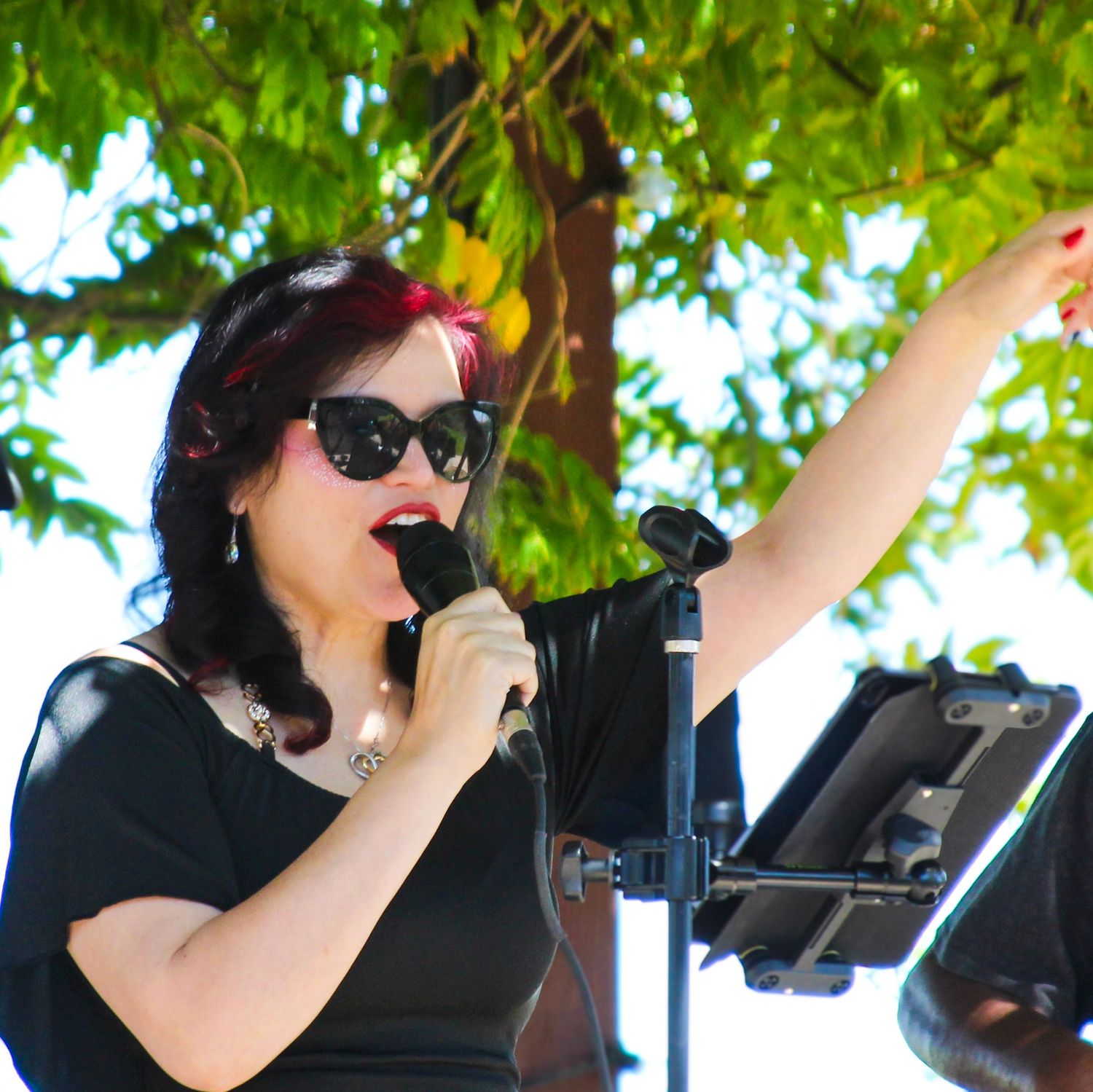 A woman in black outfit and sunglasses sings into a microphone outdoors under a green tree at a live event.