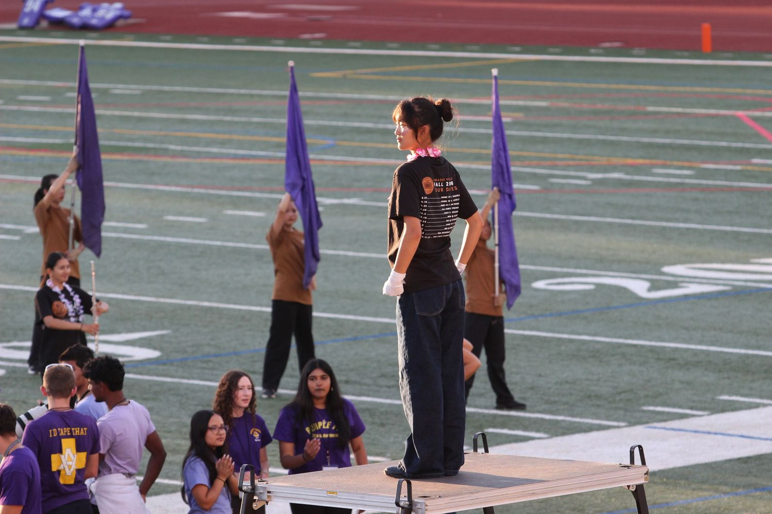 Marching band color guard members perform with purple flags on a football field during a competition.