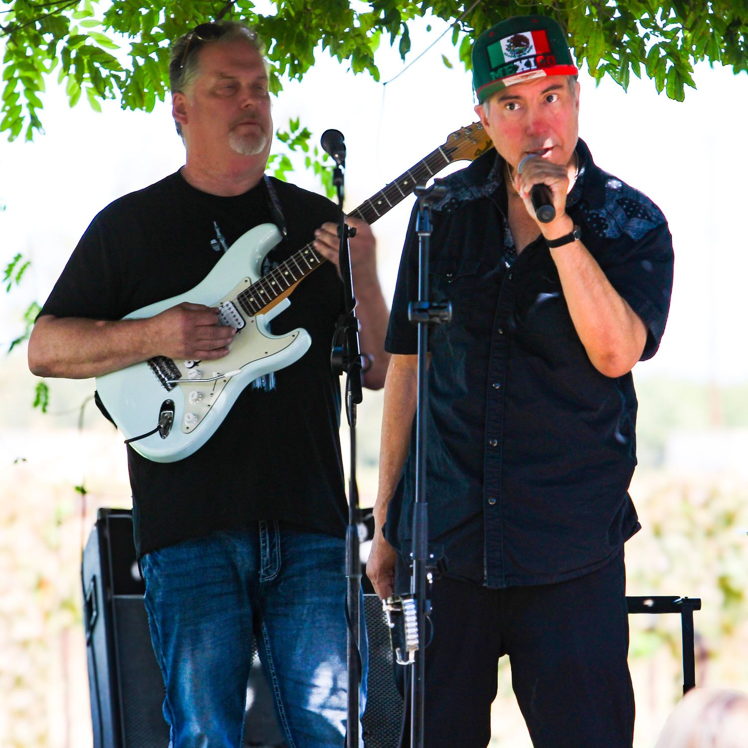Two musicians perform outdoors, one playing white electric guitar and one singing into microphone at a sunny live music event.