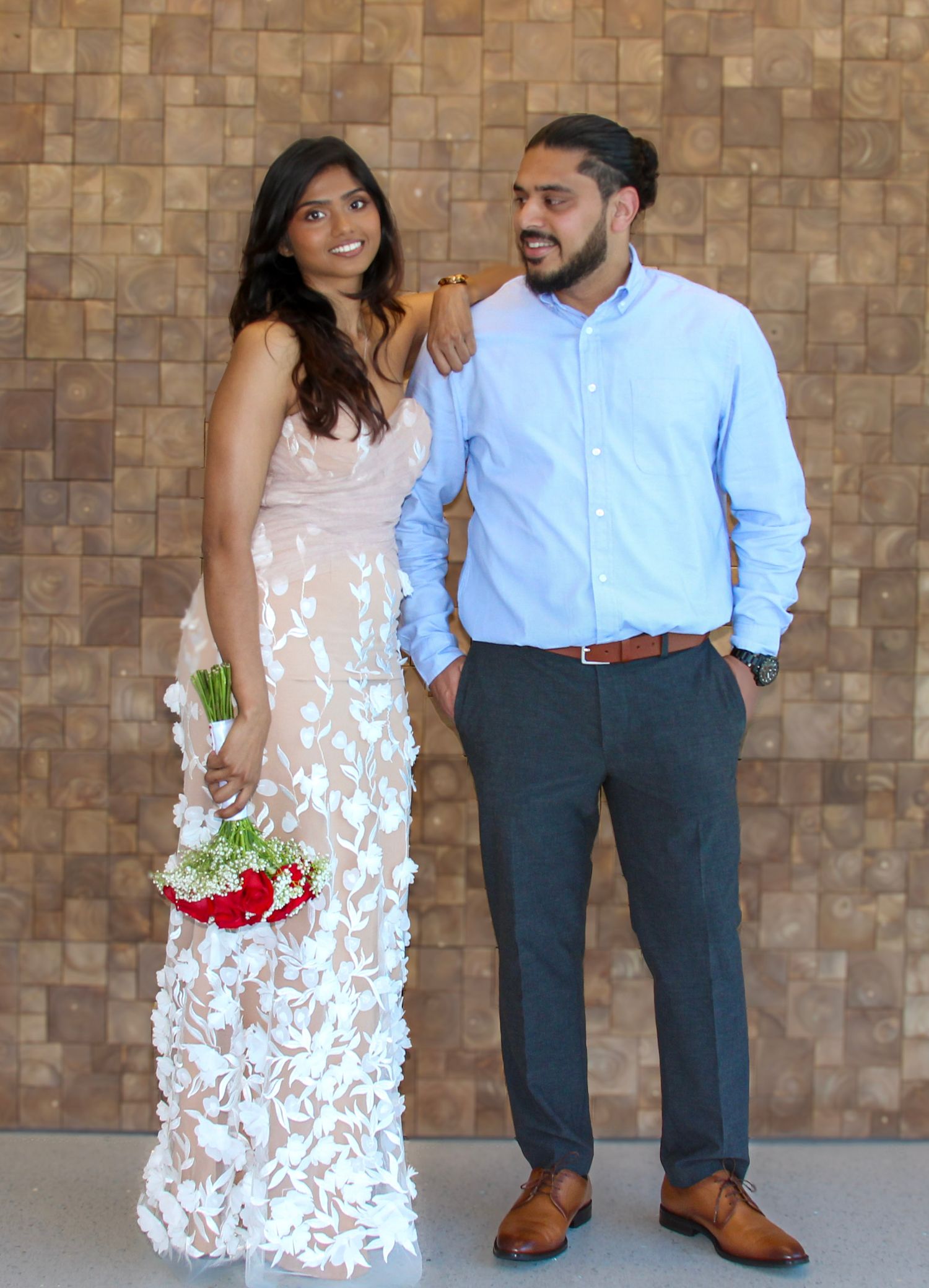 Couple posing formally, woman in white floral gown holding red bouquet, man in light blue shirt and gray pants.