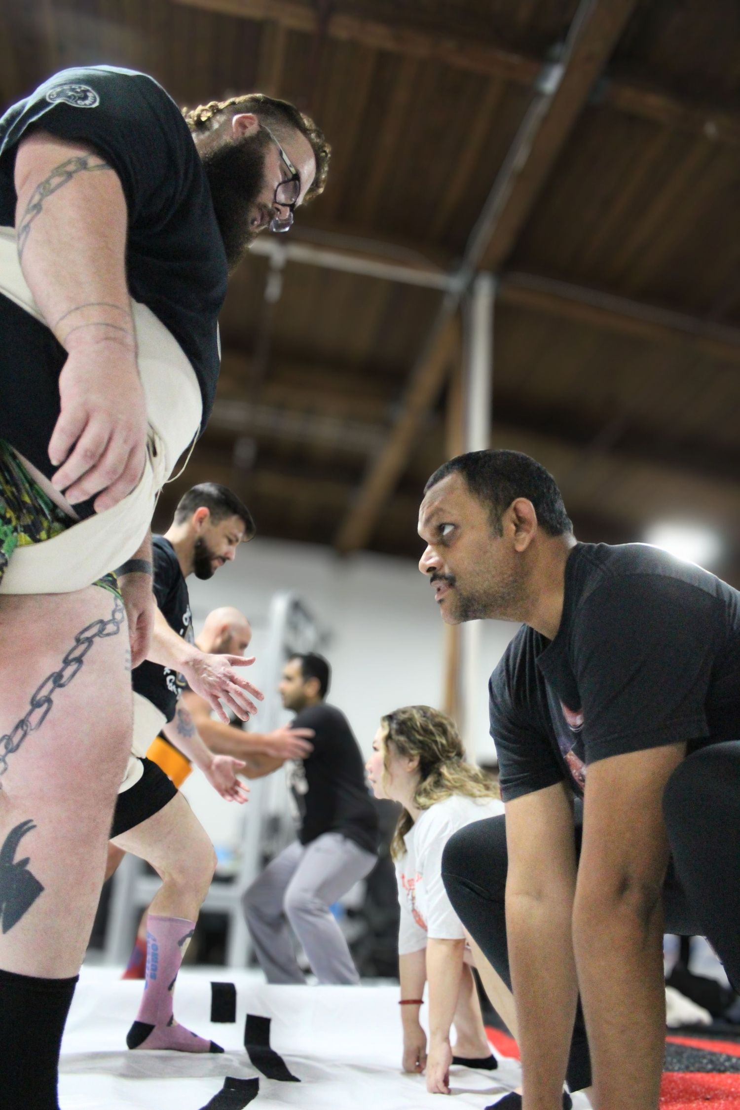 Two men in a martial arts gym, one tattooed with knee brace, another in black shirt, during a grappling training session.