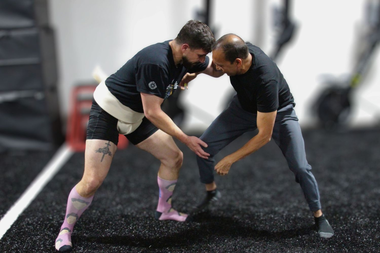 Two athletes practice wrestling techniques on black rubber flooring in a modern gym facility.
