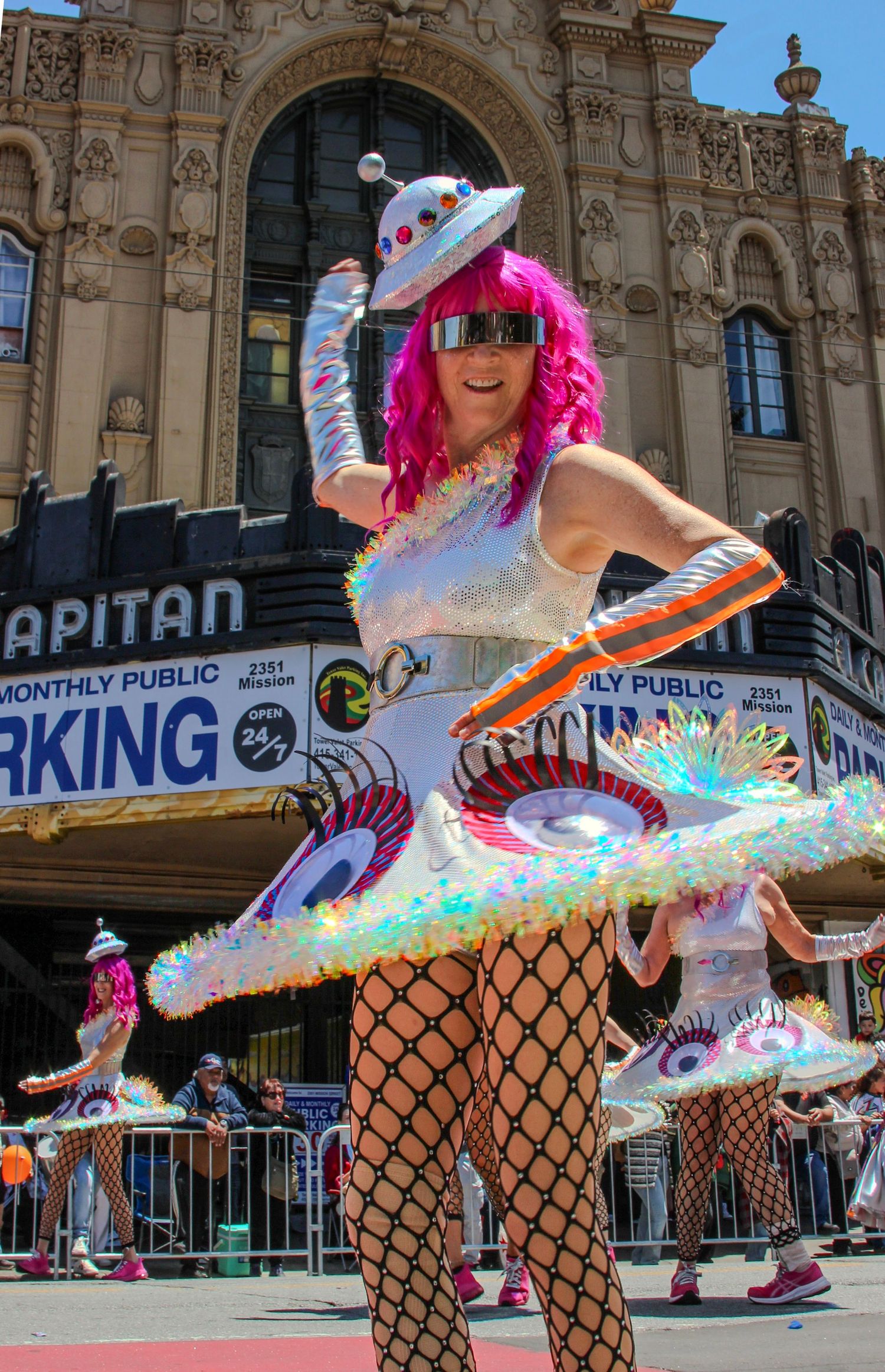 Performer in silver futuristic costume with pink wig and iridescent skirt dancing at a street parade near El Capitan Theatre.