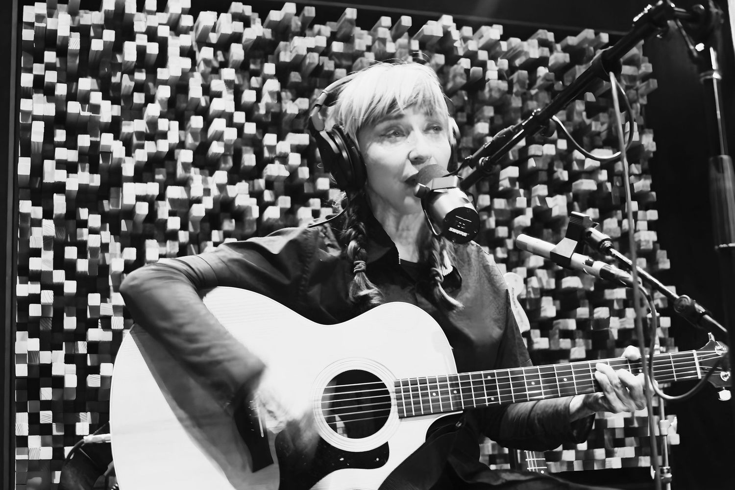 Black and white photo of a woman with braids singing into a microphone while playing acoustic guitar in a studio.
