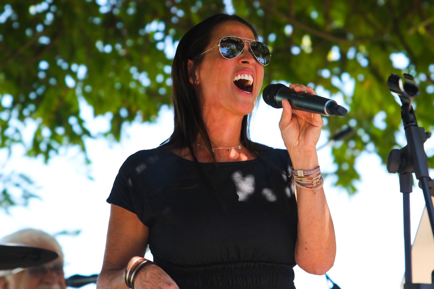 Female vocalist singing into a microphone wearing sunglasses at an outdoor concert.