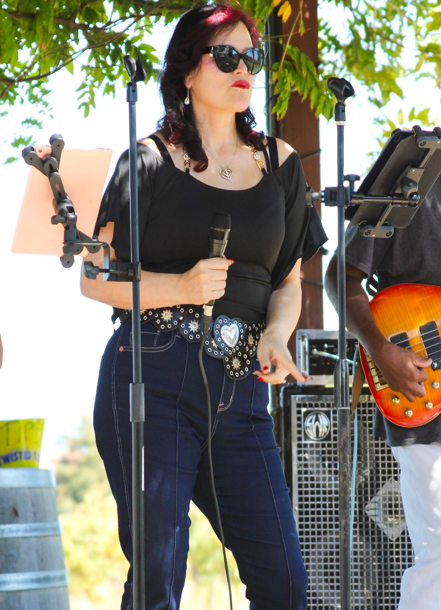 A female vocalist in black top, jeans, and sunglasses performs on an outdoor stage holding a microphone.