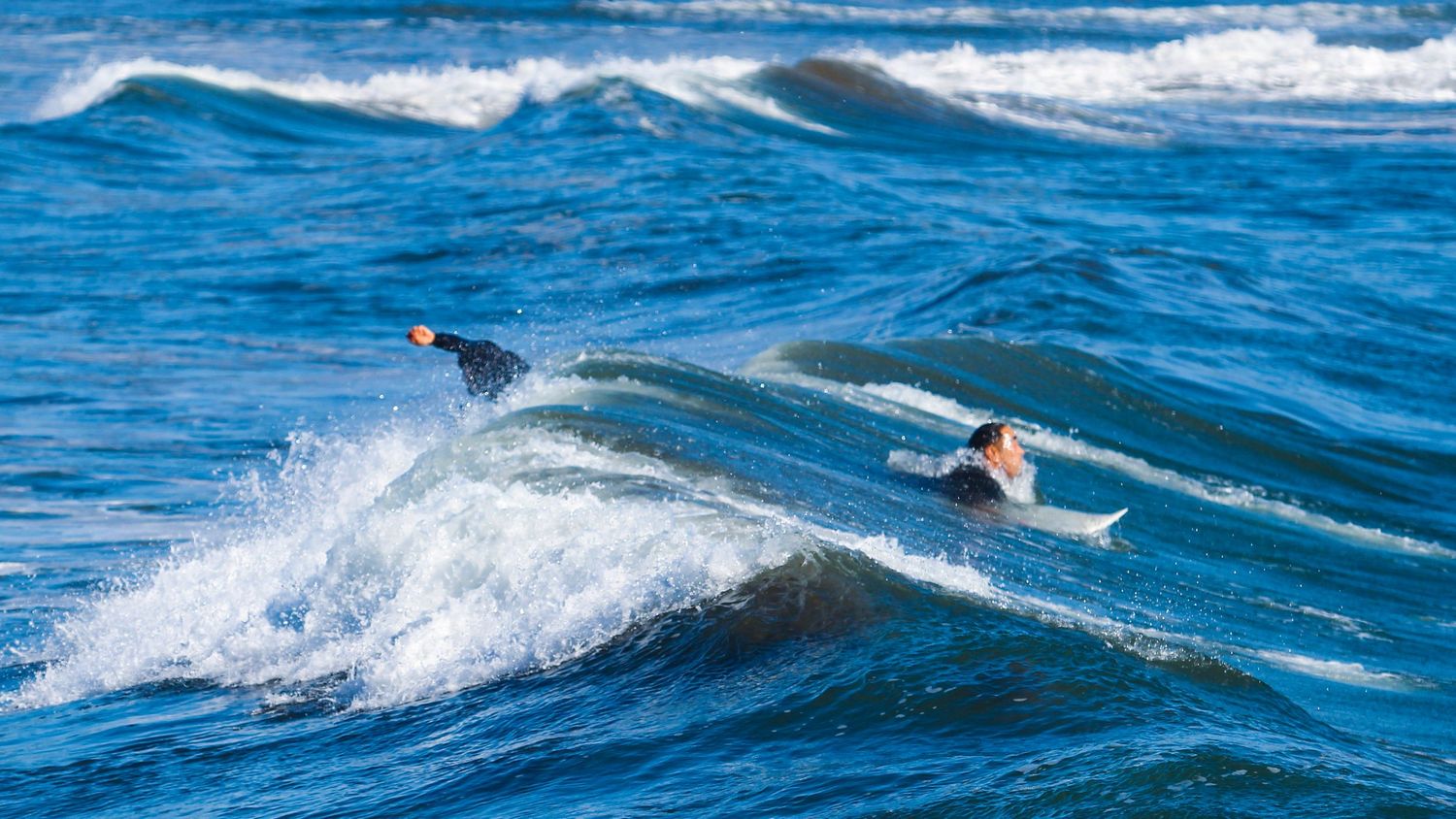 Two surfers riding ocean waves in blue water, one wiping out while the other catches a wave.
